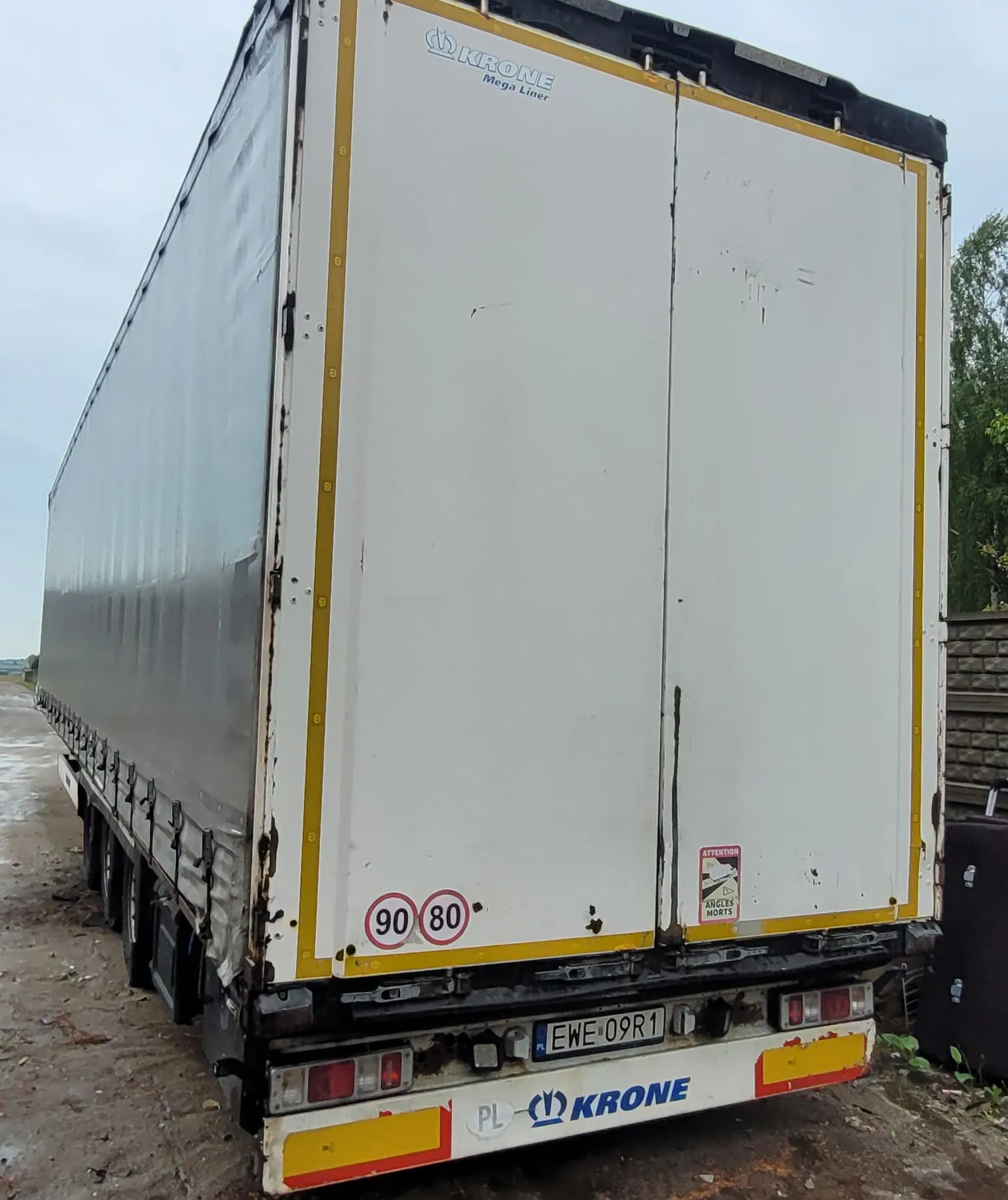 Zdjęcie tyłu i boku białej naczepy ciężarówki marki Krone Mega Liner. Na tylnych drzwiach widoczne są okrągłe znaki ograniczenia prędkości '90' i '80' oraz czerwona naklejka ostrzegawcza 'ATTENTION ANGLES MORTS'. Na dolnej części naczepy znajduje się tablica rejestracyjna 'EWE 09R1' oraz napis 'PL KRONE'. Bok naczepy jest wykonany z ciemnoszarego materiału plandekowego.
