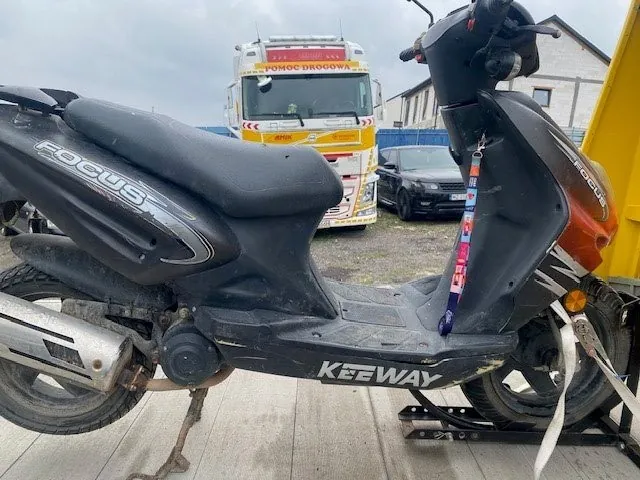 Czarny skuter marki FOCUS z napisem KEEWAY na dolnej części, zaparkowany na betonowej powierzchni. Na kierownicy wiszą kolorowe breloczki. W tle żółto-biały ciężarówka z napisem POMOC DROGOWA, czarny samochód osobowy oraz budynek z ceglaną elewacją. Niebo jest zachmurzone.