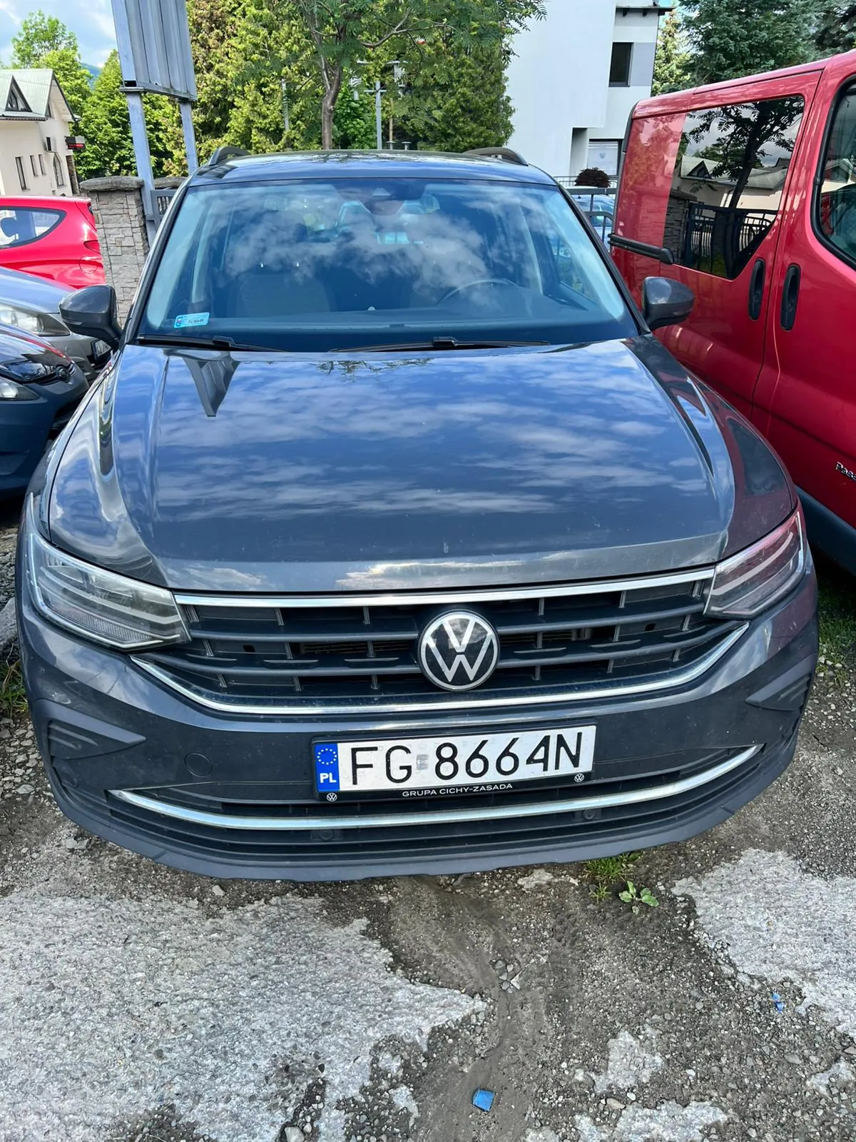 Szary samochód marki Volkswagen zaparkowany przodem do obiektywu. Na masce i przedniej szybie odbijają się chmury. Samochód posiada polską tablicę rejestracyjną o numerze FG 8664N. Poniżej tablicy znajduje się napis GRUPA CICHY-ZASADA. Po prawej stronie widoczny jest fragment czerwonego vana, a po lewej fragment innego czerwonego i ciemnego samochodu.