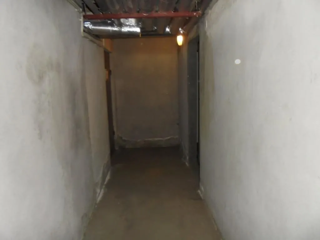 Wąski korytarz z betonowymi ścianami i podłogą. Na suficie widoczne są metalowe rury i kanały wentylacyjne. Po prawej stronie znajduje się wisząca lampa oświetlająca przestrzeń. Na końcu korytarza betonowa ściana, a po prawej stronie częściowo widoczne drzwi. Ściany mają ślady zużycia i plamy.