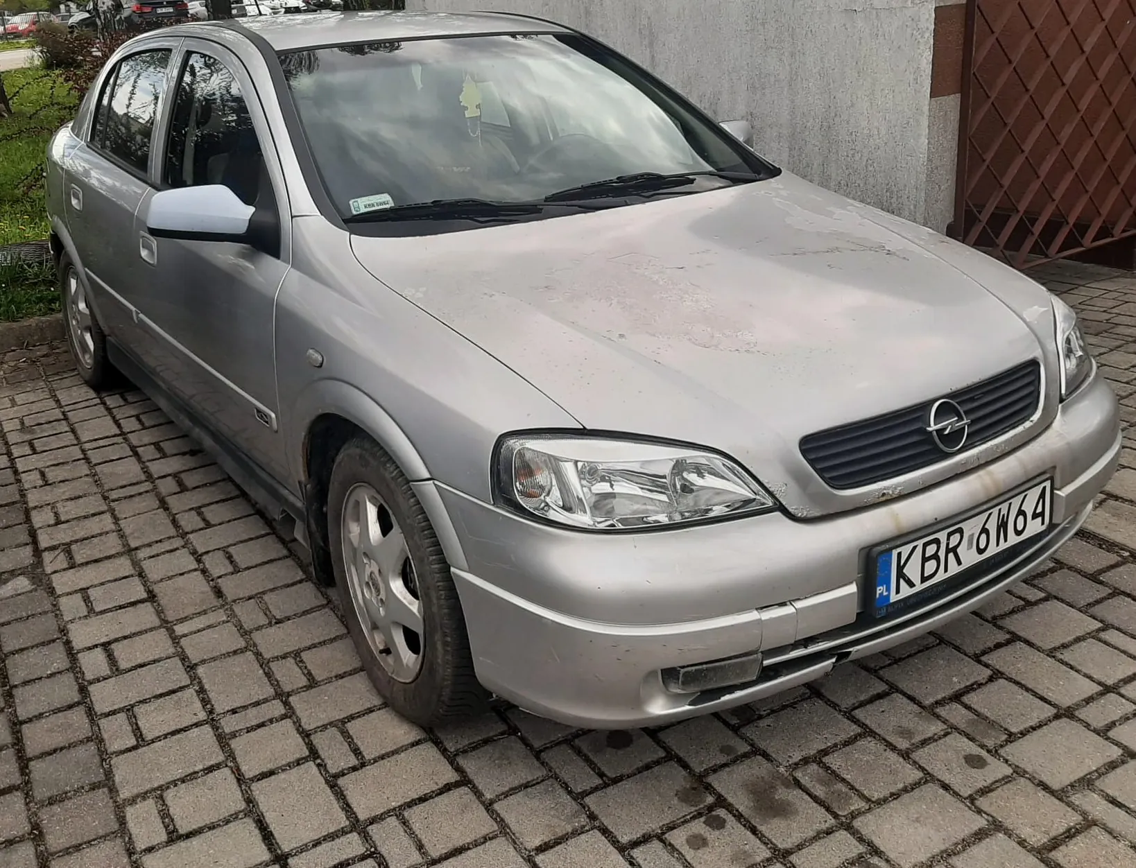 Srebrny samochód marki Opel zaparkowany na brukowanej powierzchni. Widoczna tablica rejestracyjna z polskim kodem PL i numerem KBR 6W64. Samochód ma zarysowane ślady brudu na karoserii, a na przedniej szybie znajduje się naklejka. W tle betonowa ściana budynku i brązowa konstrukcja z siatką. Po lewej stronie zielona trawa i krzewy.