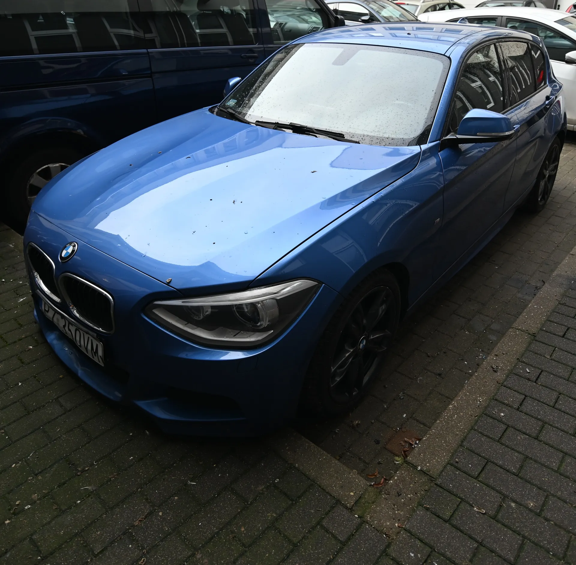 Niebieski samochód BMW z tablicą rejestracyjną P7 250VM zaparkowany na brukowanej powierzchni. Przednią szybę zasłania parasol przeciwsłoneczny. W tle widoczne inne pojazdy, w tym ciemnoniebieski van i biały samochód. Na karoserii ślady brudu.