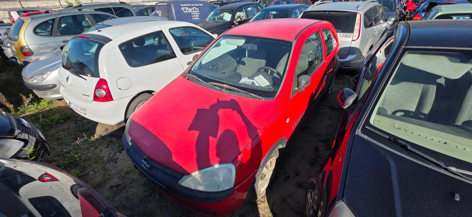 Rząd zaparkowanych samochodów osobowych na placu. Na pierwszym planie znajduje się czerwony, trzydrzwiowy samochód marki Opel Corsa. Obok niego stoi biały samochód marki Renault Scenic z tablicą rejestracyjną P 491 AH. Po prawej stronie widoczny jest fragment czarnego samochodu, a w tle kolejne pojazdy w kolorach srebrnym, niebieskim i czarnym. Na masce czerwonego samochodu widoczny jest wyraźny cień osoby robiącej zdjęcie.