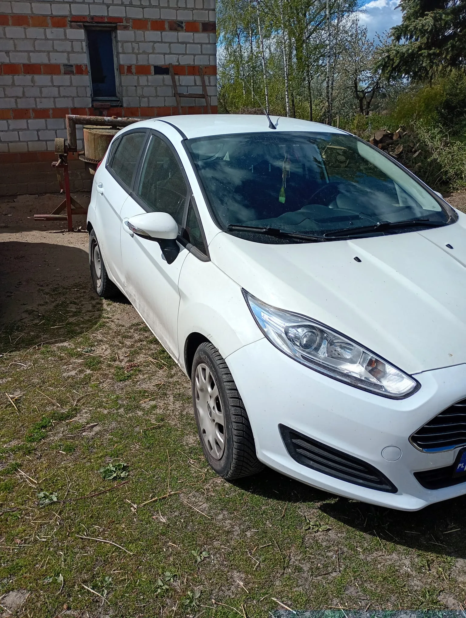 Biały samochód osobowy marki Ford zaparkowany na trawiastym terenie z widocznymi plamami ziemi. W tle budynek z cegły i bloczków z czerwonymi pasami, z oknem i drabiną opartą o ścianę. Po prawej stronie drzewa, krzewy i stos drewna. Na tablicy rejestracyjnej widoczny fragment 'PL'. Niebo częściowo zachmurzone. Przed samochodem znajduje się metalowa konstrukcja z beczką.