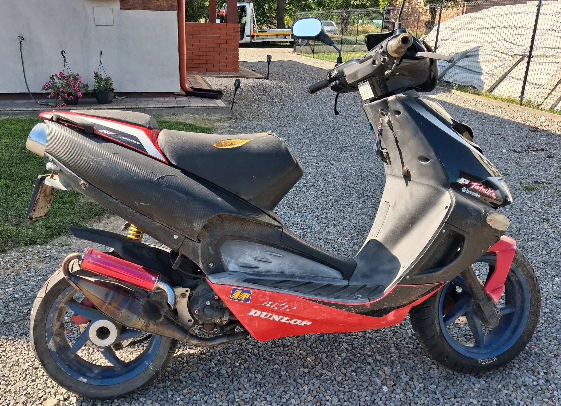 Czarny skuter z czerwonymi elementami nadwozia, zaparkowany na żwirowym podjeździe. Na boku skutera widnieje napis 'DUNLOP' oraz logo 'iR'. Pojazd ma czarne felgi i srebrny wydech. W tle widać fragment białego budynku, ceglany murek oraz zieloną roślinność.
