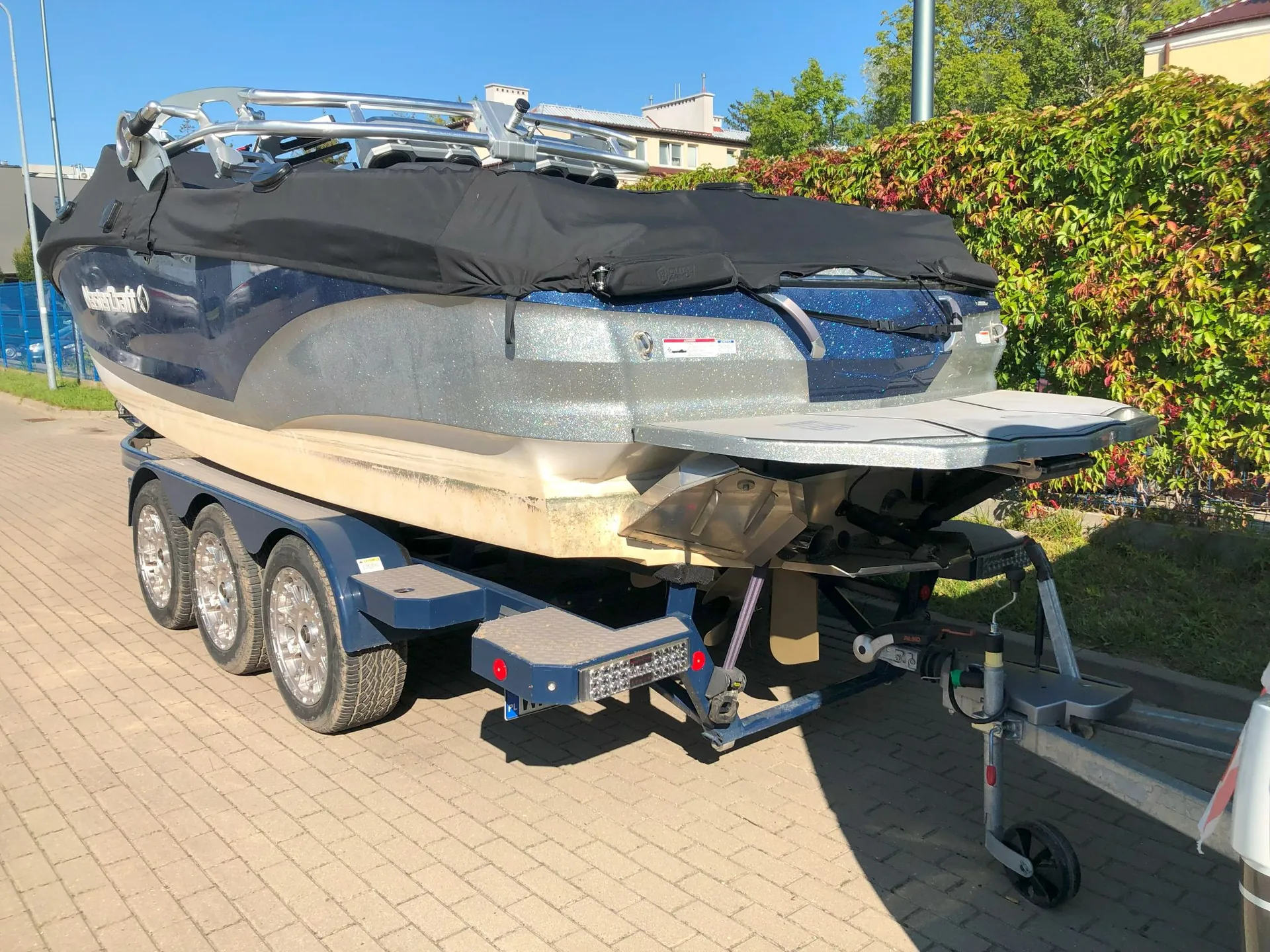 Motorówka w kolorze niebieskim z brokatowym wykończeniem, przykryta czarną plandeką, zaparkowana na dwuosiowej przyczepie transportowej na kostce brukowej. Łódź posiada srebrne ramy nad kokpitem oraz platformę kąpielową z tyłu. W tle widoczne są zielone krzewy i fragmenty budynków pod bezchmurnym, niebieskim niebem.