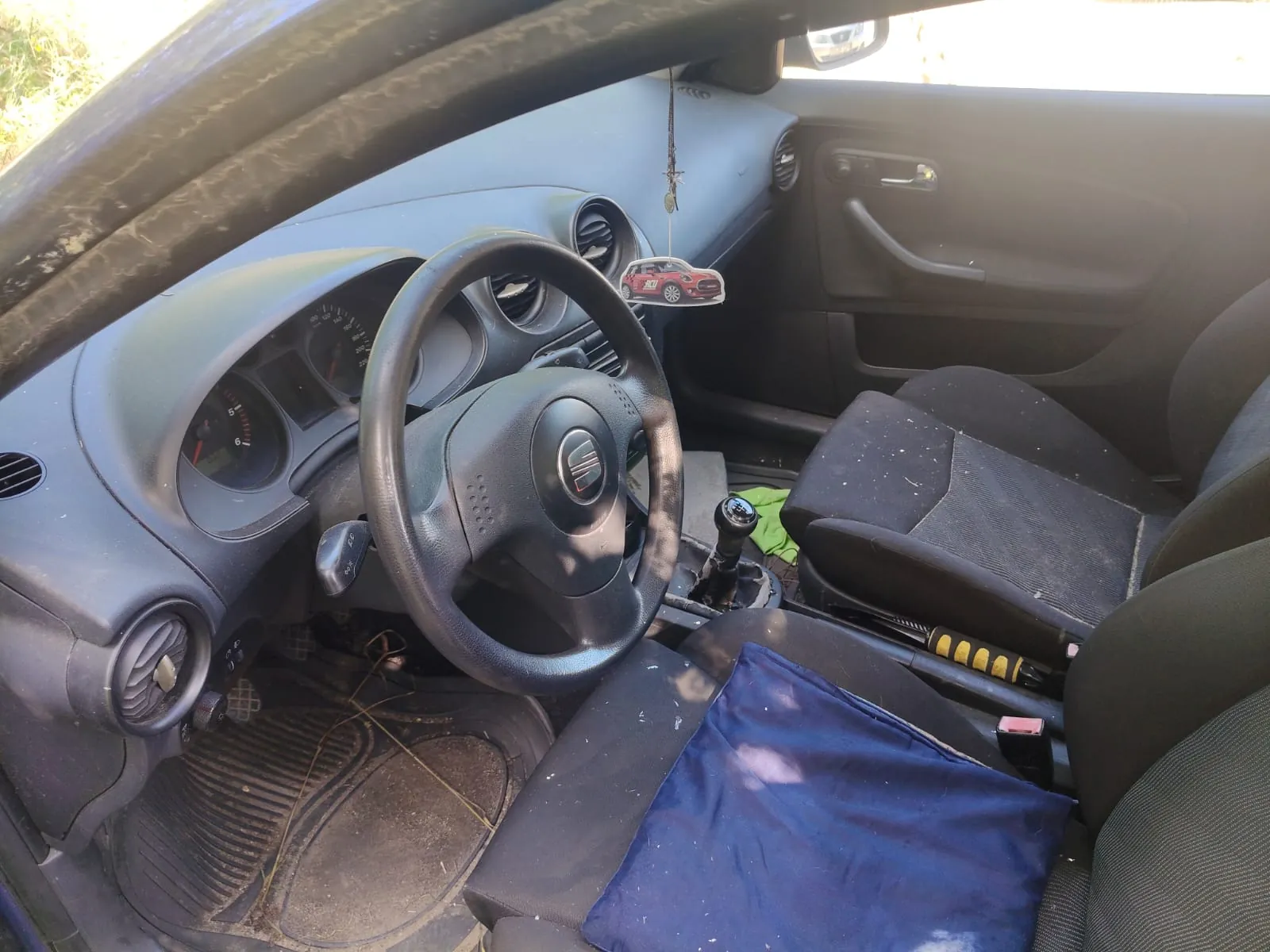 Wnętrze samochodu z perspektywy kierowcy. Widoczna jest czarna kierownica z logo marki Seat, deska rozdzielcza z analogowymi wskaźnikami prędkości i obrotów oraz dwa przednie fotele z ciemnej tkaniny. Na fotelu pasażera leży niebieska szmatka. Na lusterku wstecznym wisi mała ozdoba w kształcie czerwonego samochodu. Podłoga wyłożona jest gumowymi dywanikami.