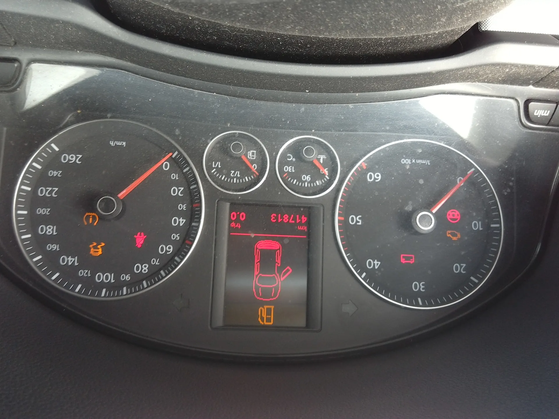 Tablica przyrządów samochodowych z czarnym tłem. Po lewej stronie prędkościomierz z zakresem 0-260 km/h, wskazówka na 0 km/h, ikony ostrzegawcze: pomarańczowy symbol informacyjny, czerwony pas bezpieczeństwa i pomarańczowy symbol zbiornika paliwa. W centrum wyświetlacz z czerwonymi cyframi: 417813 (przebieg), 0.0 (trip), ikoną samochodu z otwartą klapą bagażnika i pomarańczowym symbolem pompy paliwowej. Po prawej stronie tachometr z zakresem 0-60 (1/min x100), wskazówka na 0, ikony ostrzegawcze: czerwony symbol silnika i pomarańczowy symbol akumulatora. Powyżej centralnego wyświetlacza dwa małe wskaźniki: poziom paliwa (1/1) i temperatura chłodniczy (90°C). W prawym górnym rogu przycisk oznaczony 'min'.