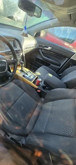 Wnętrze samochodu marki Audi z czarnymi siedzeniami w geometryczny wzór, kierownicą z logo Audi, centralną konsolą z przyciskami i dźwignią biegów. W uchwycie na napoje znajduje się czerwona puszka. Widoczne są przednie siedzenia, lusterko wsteczne oraz fragment czerwonego samochodu za oknem.