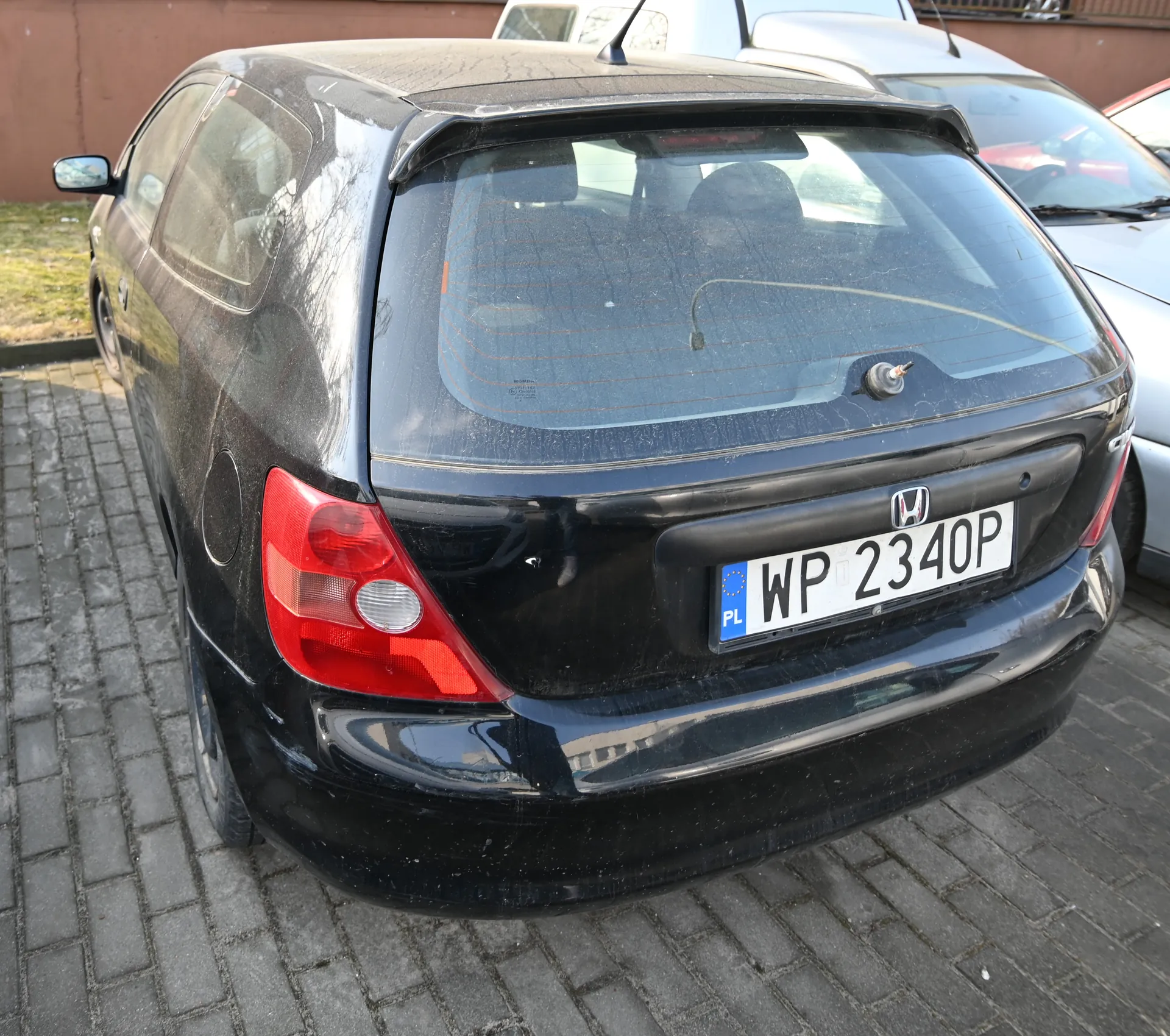 Czarny samochód marki Honda widziany od tyłu, zaparkowany na brukowanej powierzchni. Widoczna tablica rejestracyjna z białym tłem, czarnym napisem 'WP 2340P' oraz niebieskim pasem z napisem 'PL' i symbolem Unii Europejskiej. W tle inne pojazdy (biały i srebrny samochód) oraz brązowa ściana budynku. Tylne światła samochodu są czerwone, a na szybie tylnej widoczne są ślady brudu.