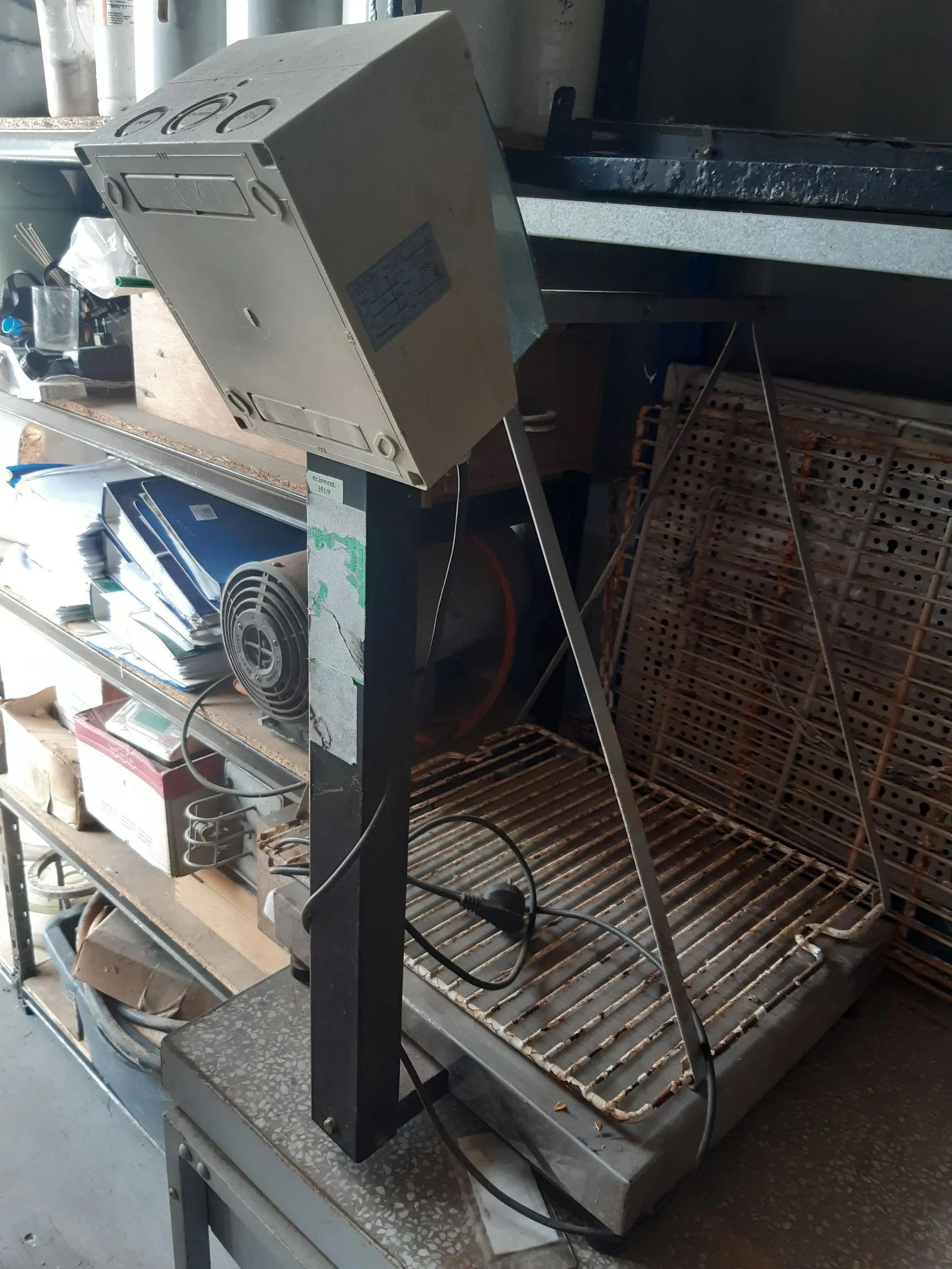 Urządzenie techniczne zamontowane na czarnym, pionowym wsporniku. Na górze znajduje się biała, plastikowa obudowa z przyciskami i etykietą techniczną. Poniżej znajduje się metalowa kratka grzewcza lub chłodząca z widocznym kablem zasilającym. Urządzenie stoi na szarym blacie w pomieszczeniu przypominającym warsztat lub magazyn, w tle widoczne są regały z dokumentami i innymi przedmiotami.