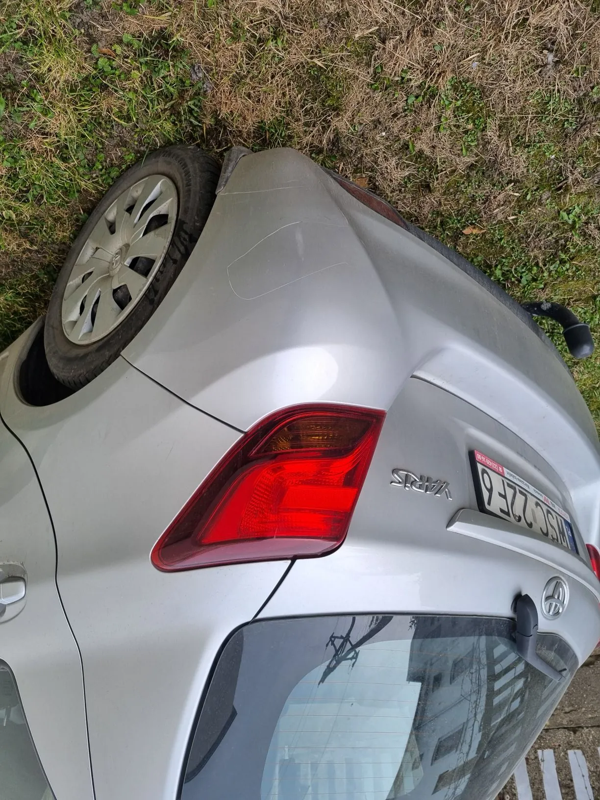 Zbliżenie na tylną część srebrnego samochodu Toyota Yaris zaparkowanego na trawie. Widoczne są czerwone światło tylnego, tablica rejestracyjna z napisem WSC 22F6, logo Toyoty oraz napis YARIS. Trawa w tle jest częściowo zielona, częściowo sucha. Na tylnej szybie odbijają się budynki i linie energetyczne.