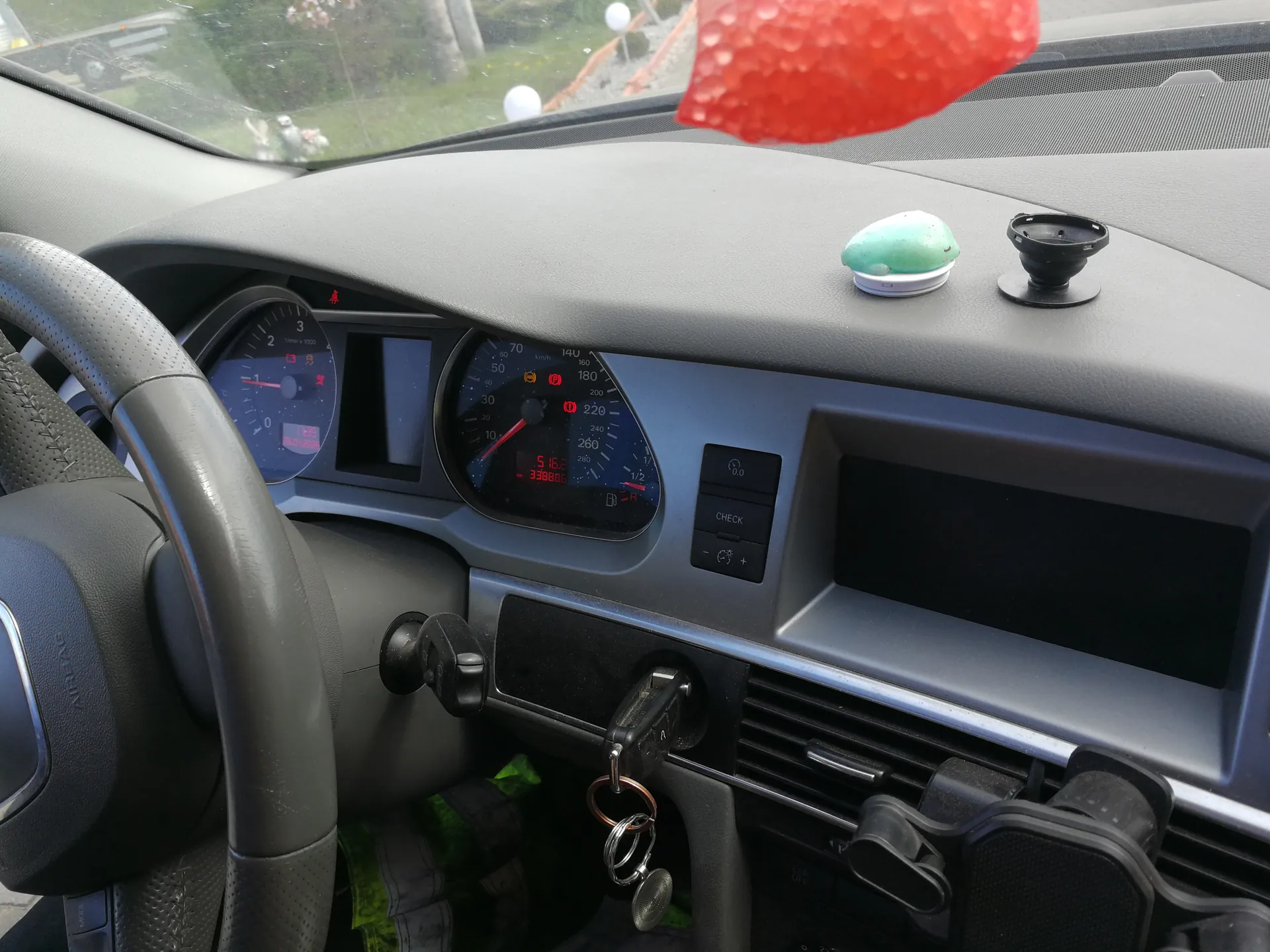Wnętrze samochodu z widoczną deską rozdzielczą, kierownicą po lewej stronie, dwoma wskaźnikami (prędkościomierz do 260 km/h i tachometr do 7000 obr./min), cyfrowym wyświetlaczem pokazującym 5:16.2 i przebieg 33886 km. Na desce rozdzielczej znajdują się zielono-biały przedmiot, czarny uchwyt na ssawce oraz czerwony, kropkowany przedmiot wiszący na lusterku wstecznym. W stacyjce znajdują się kluczyki z brelokami. Widoczne są również przyciski oznaczone 'CHECK' oraz wentylacyjne otwory. Przez przednią szybę widać zieloną trawę i białe kule na zewnątrz.