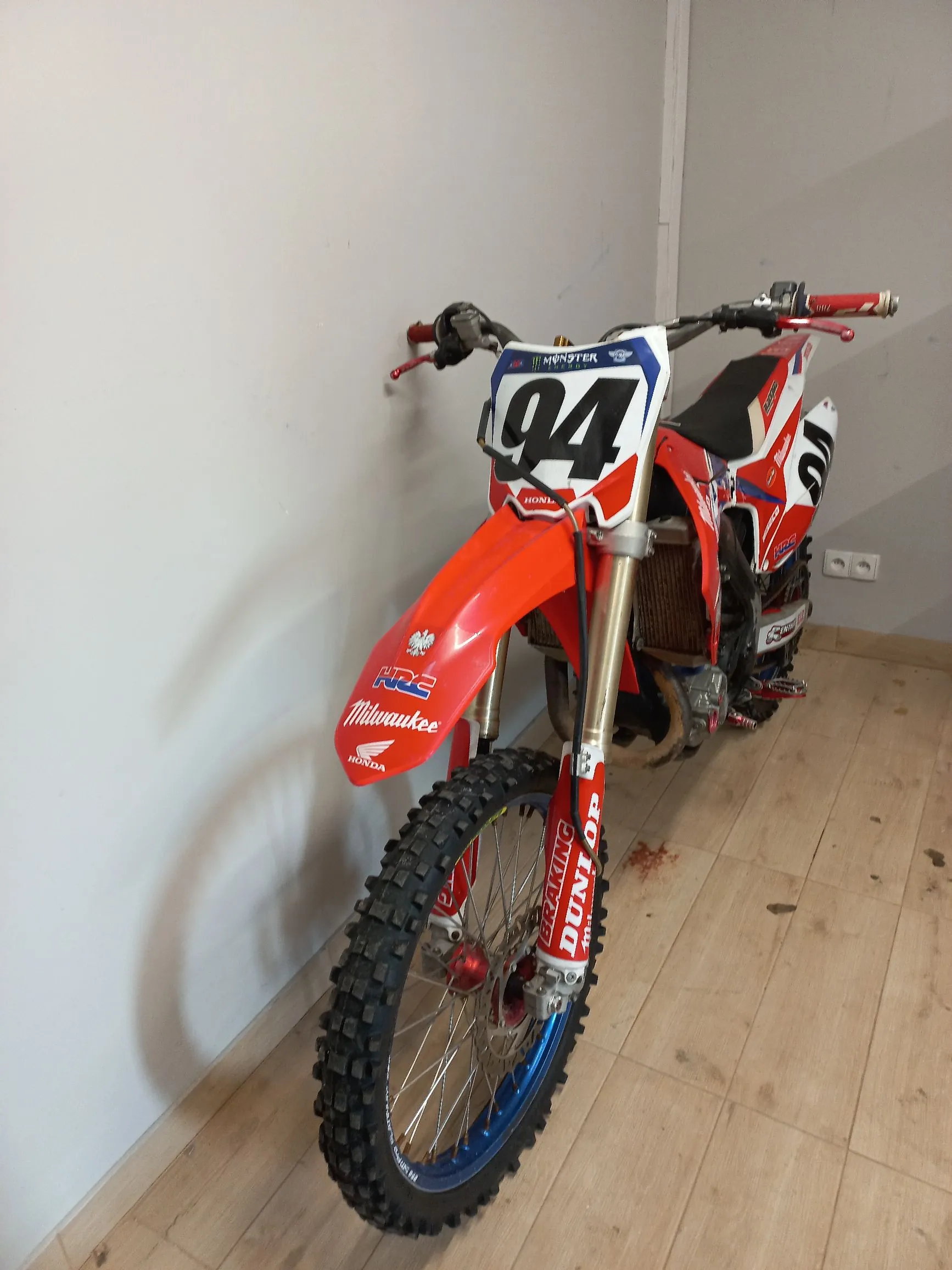 Czerwony motocykl crossowy marki Honda z numerem 94 na przedniej owiewce. Motocykl posiada liczne naklejki sponsorów, w tym Milwaukee i Dunlop. Stoi on na drewnianej podłodze w jasnym pomieszczeniu przy białej ścianie.