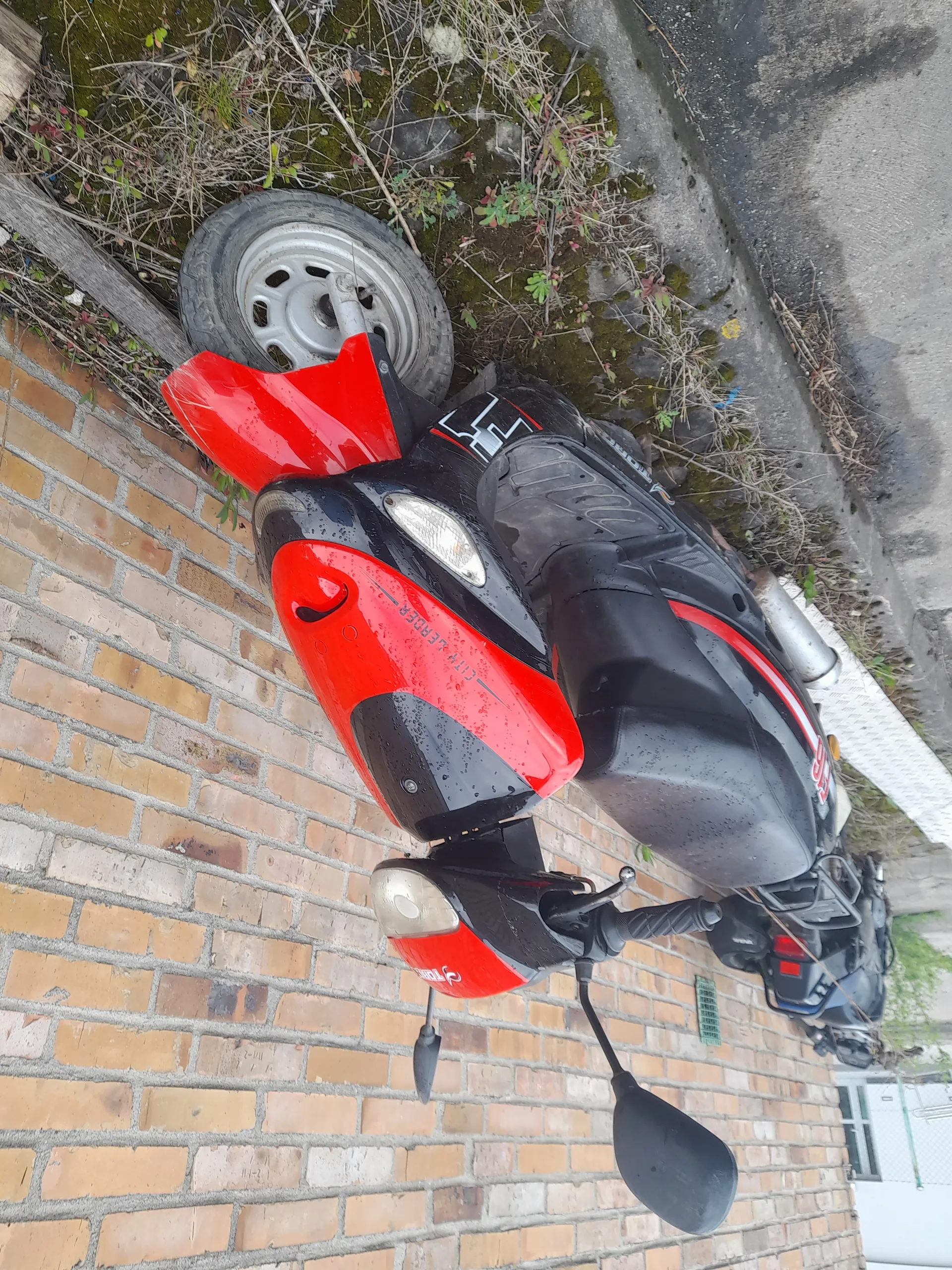 Czerwono-czarny skuter zaparkowany przy ceglanej ścianie. Na skuterze widoczny napis 'CITY LEADER' oraz grafika z cyfrą '50'. Skuter posiada dwa lusterka, reflektory i jest umieszczony na krawężniku z trawą i mchem. W tle ceglana ściana oraz fragment drogi.