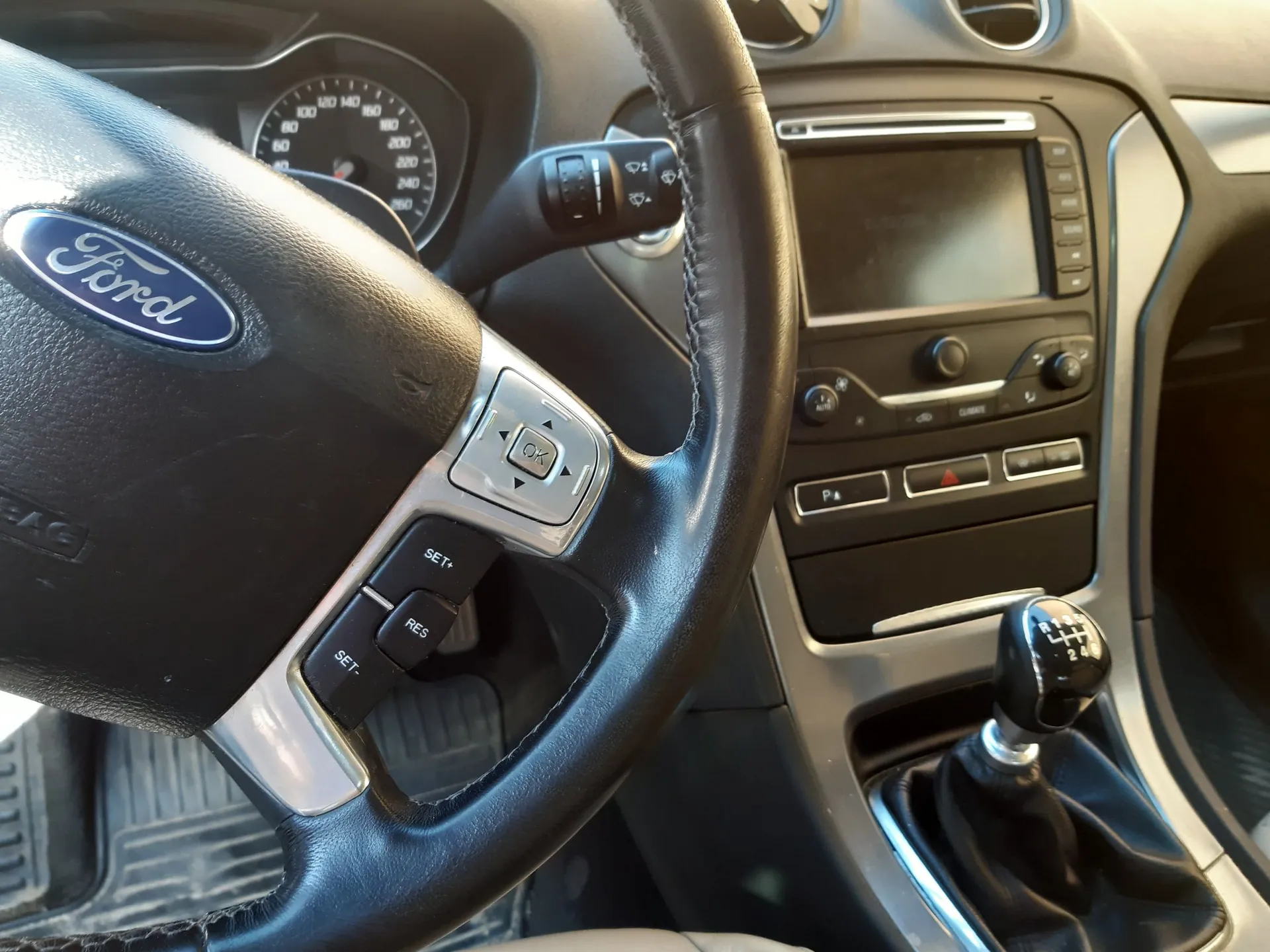 Wnętrze samochodu marki Ford z perspektywy kierowcy. Widoczna jest czarna skórzana kierownica z logo Ford na środku oraz przyciskami sterującymi po prawej stronie. W tle znajduje się deska rozdzielcza z analogowym prędkościomierzem, ekranem multimedialnym, przyciskami klimatyzacji oraz lewarkiem zmiany biegów w manualnej skrzyni przekładni.