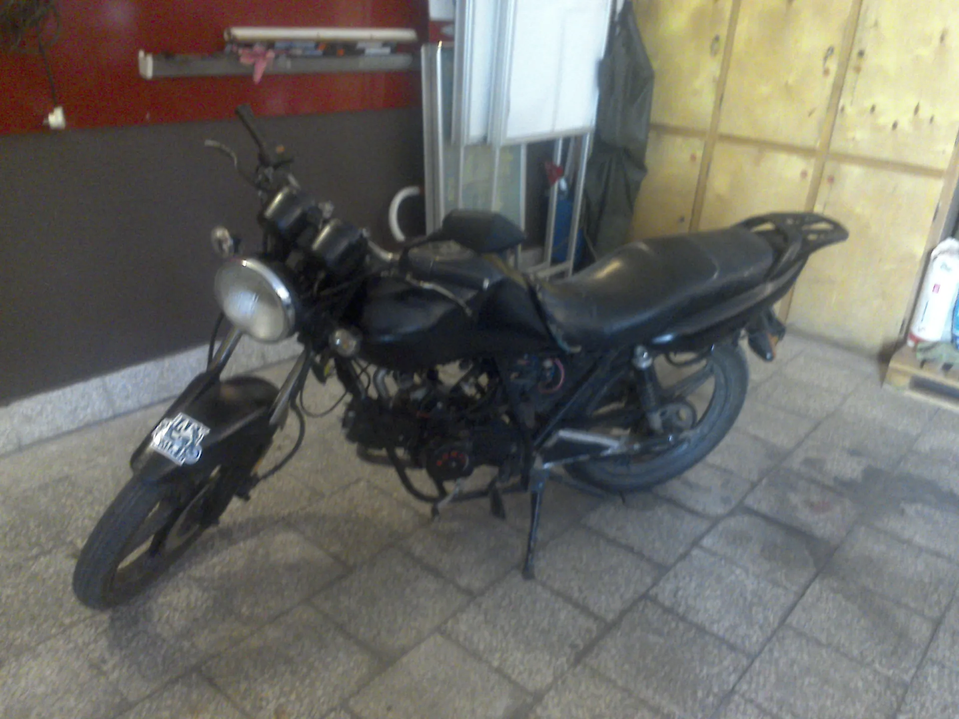 Czarny motocykl zaparkowany w pomieszczeniu na szarej, płytkowej podłodze. Pojazd jest ustawiony bokiem, z przodu widoczna jest okrągła lampa, a w tle znajdują się ściany w kolorach czerwonym i szarym oraz drewniane panele.