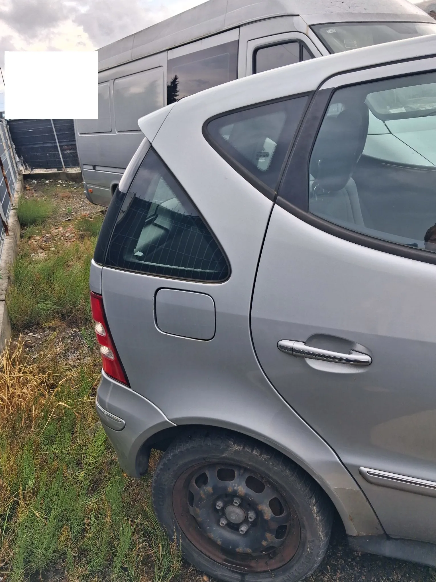 Zbliżenie na tylną, prawą część srebrnego samochodu osobowego zaparkowanego na trawiastym terenie. Widoczna jest tylna lampa, klapka wlewu paliwa, klamka drzwi oraz czarne, stalowe koło. W tle znajduje się srebrny samochód dostawczy typu bus.