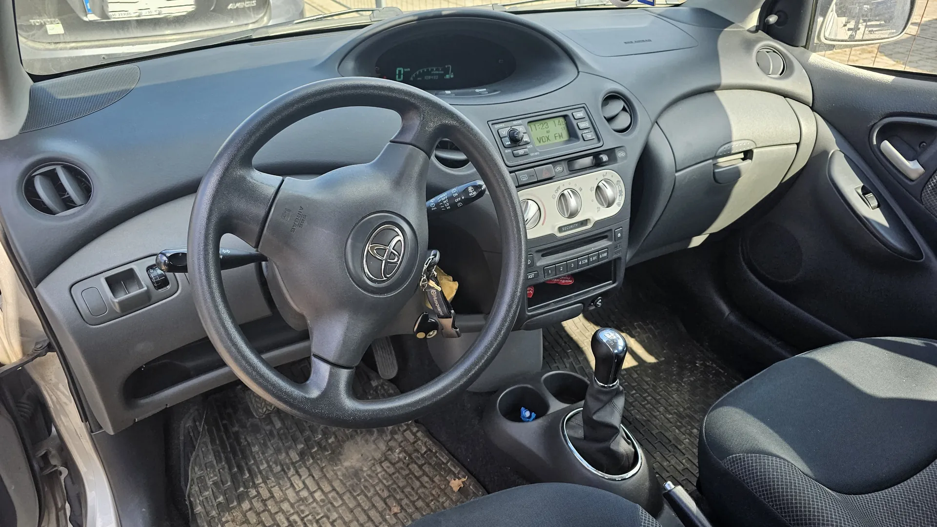 Wnętrze samochodu marki Toyota z widocznym kierownicą z logo producenta, kluczami w zapłonie, deska rozdzielcza z cyfrowym wyświetlaczem pokazującym godzinę 11:23, temperaturę 14°C oraz napisem VOX FM. W centralnej konsoli znajdują się przyciski sterujące, pokrętła klimatyzacji, dźwignia zmiany biegów oraz uchwyty na napoje. Widoczne są czarne siedzenia z wzorzystą tapicerką, podłoga z matami oraz otwarte drzwi po lewej stronie. Deska rozdzielcza i elementy są w kolorze szarym.