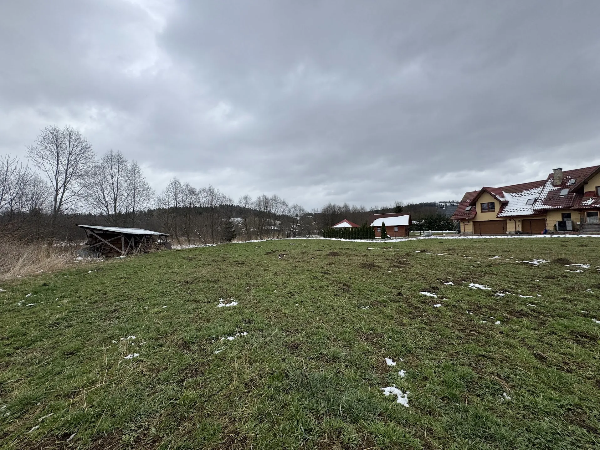 Pola trawiaste z resztkami śniegu, na tle domy z czerwonymi dachami częściowo pokrytymi śniegiem. Po lewej stronie drewniana konstrukcja z metalowym dachem, w tle drzewa bez liści i zalesiony teren. Niebo zachmurzone.