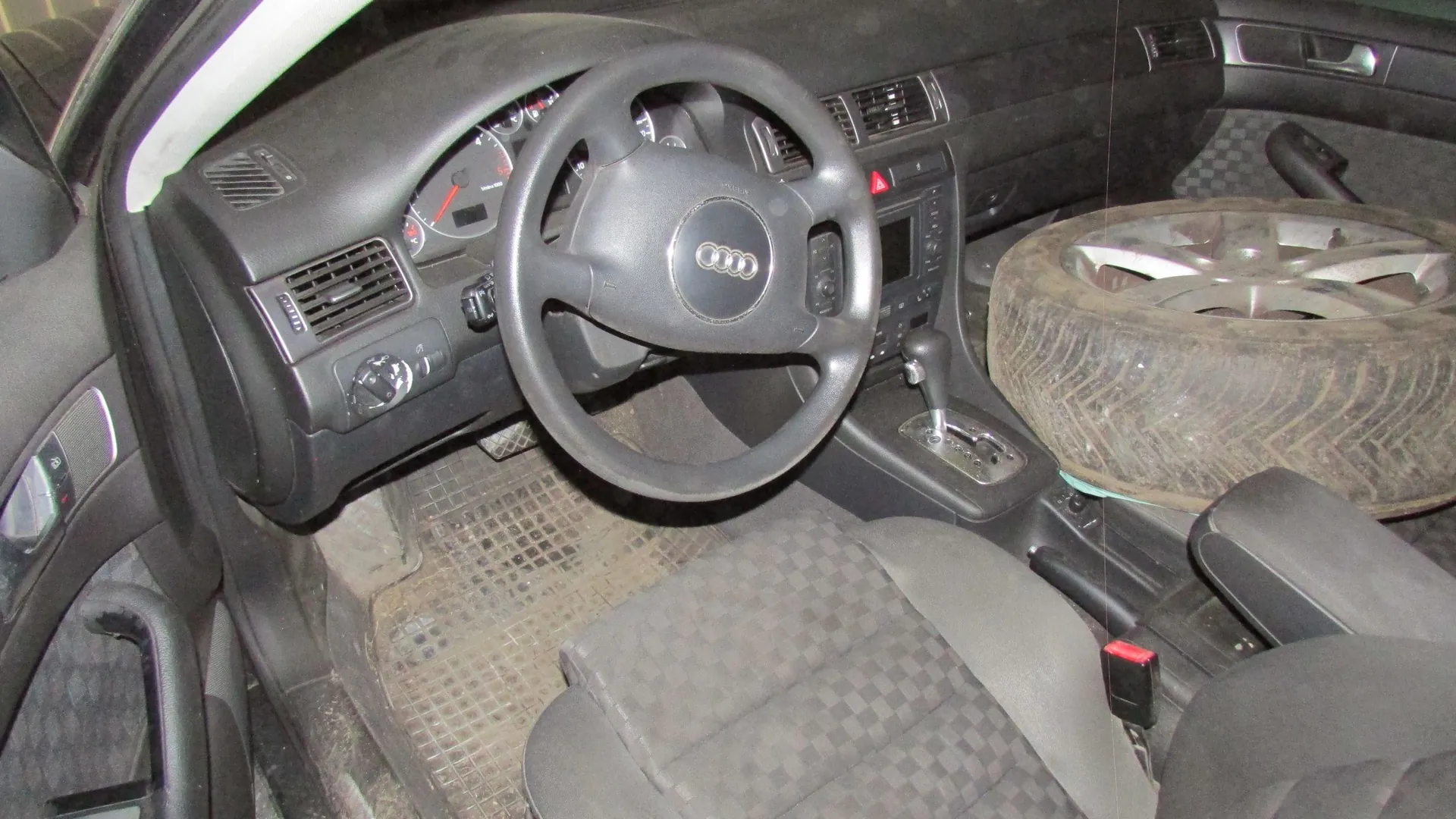 Wnętrze samochodu marki Audi z widokiem na deskę rozdzielczą, kierownicę z logo producenta oraz przedni fotel kierowcy. Na siedzeniu pasażera leży brudna opona z felgą aluminiową.