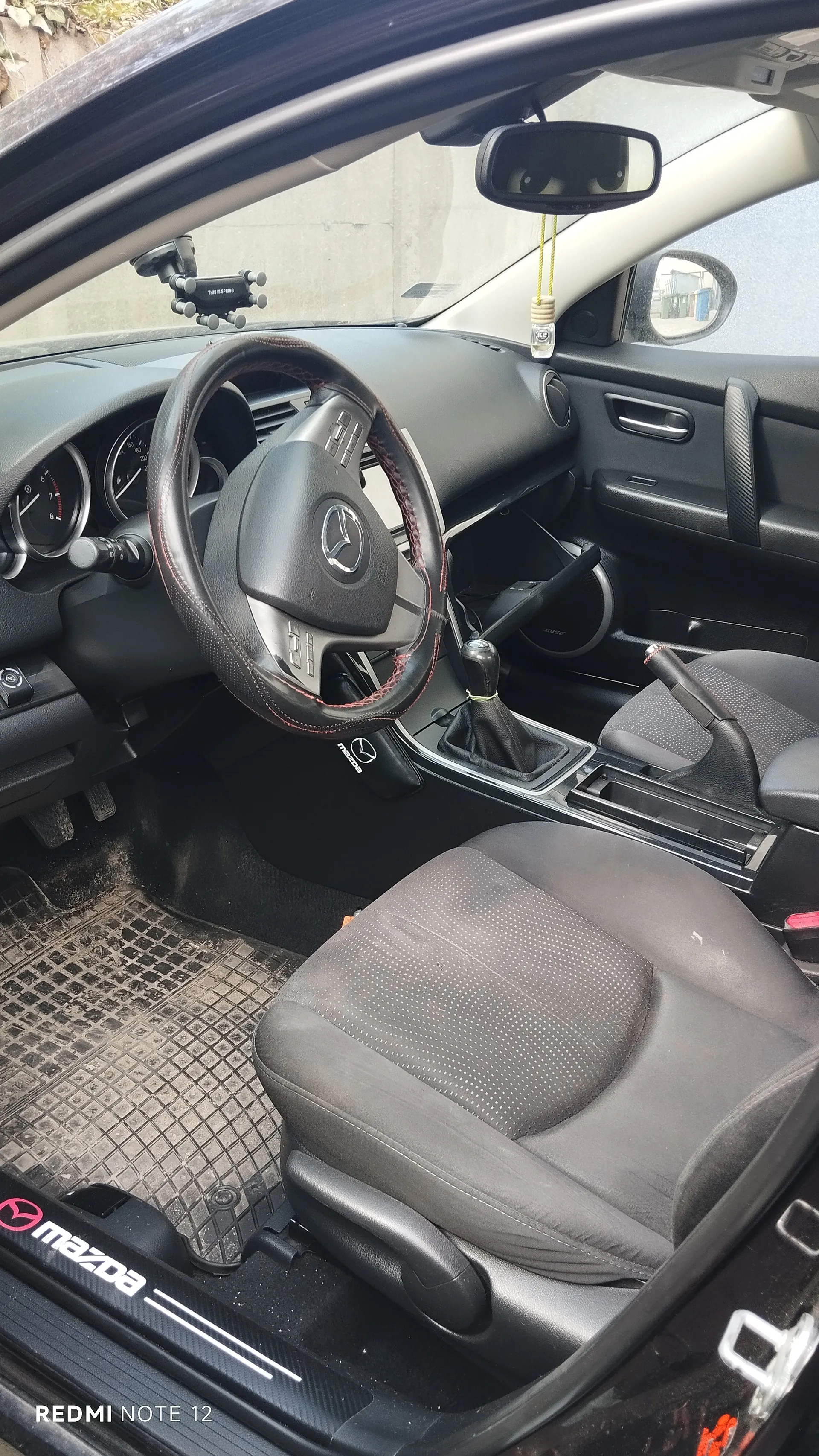 Wnętrze samochodu marki Mazda z czarną kierownicą z czerwonym przeszyciem i logo Mazdy. Widoczne są czarne i szare siedzenia z kropkowanym wzorem, deska rozdzielcza z licznikami, dźwignia zmiany biegów oraz dywaniki z napisem 'mazda'. W lusterku wstecznym i na desce rozdzielczej umocowany jest uchwyt na telefon. Na dole zdjęcia widoczny napis 'REDMI NOTE 12'.