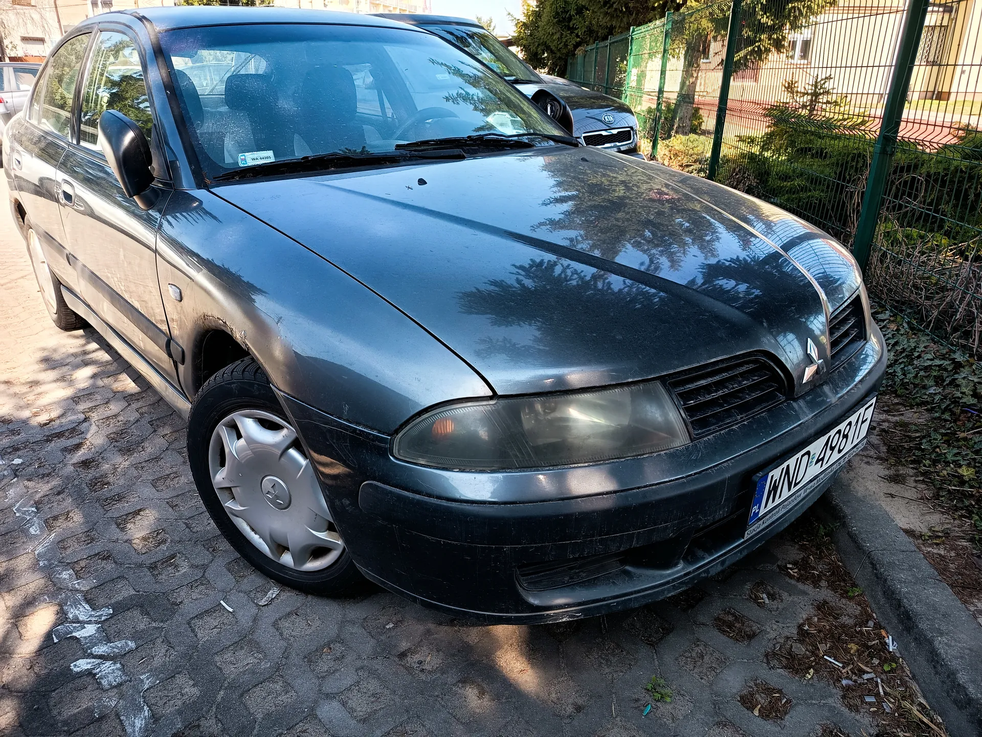 Ciemnoszary samochód osobowy marki Mitsubishi zaparkowany na brukowanej ulicy. Widoczna tablica rejestracyjna z napisem WND 4981F oraz niebieskim pasem po lewej stronie. W tle drugi samochód, zielone ogrodzenie metalowe, krzewy i budynki. Słoneczny dzień, cienie na jezdni.