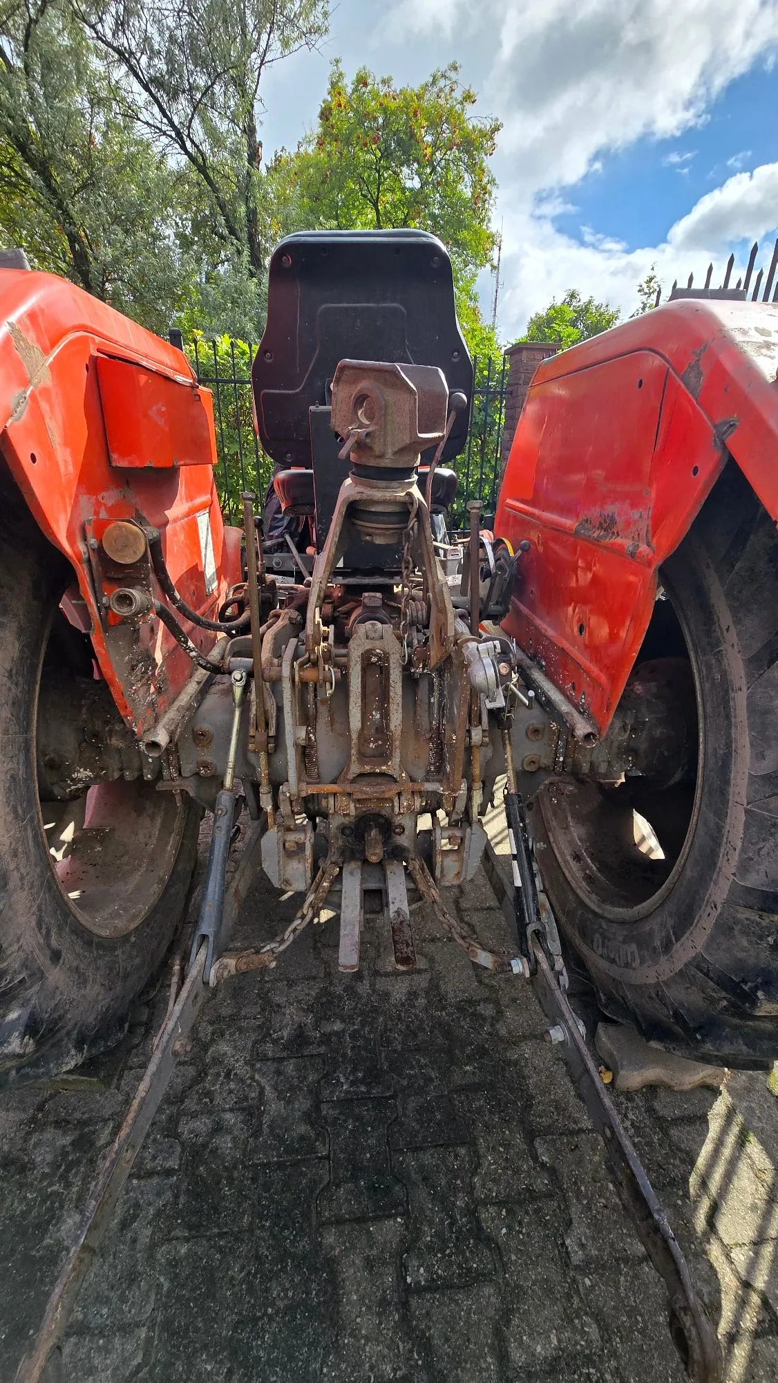 Widok z tyłu na czerwony traktor rolniczy zaparkowany na kostce brukowej. Widoczne są duże, czarne opony z obu stron, siedzenie kierowcy oraz tylny układ hydrauliczny i mechanizmy przyczepne z widocznymi śladami rdzy i zużycia. W tle znajdują się zielone drzewa i niebo z białymi chmurami.