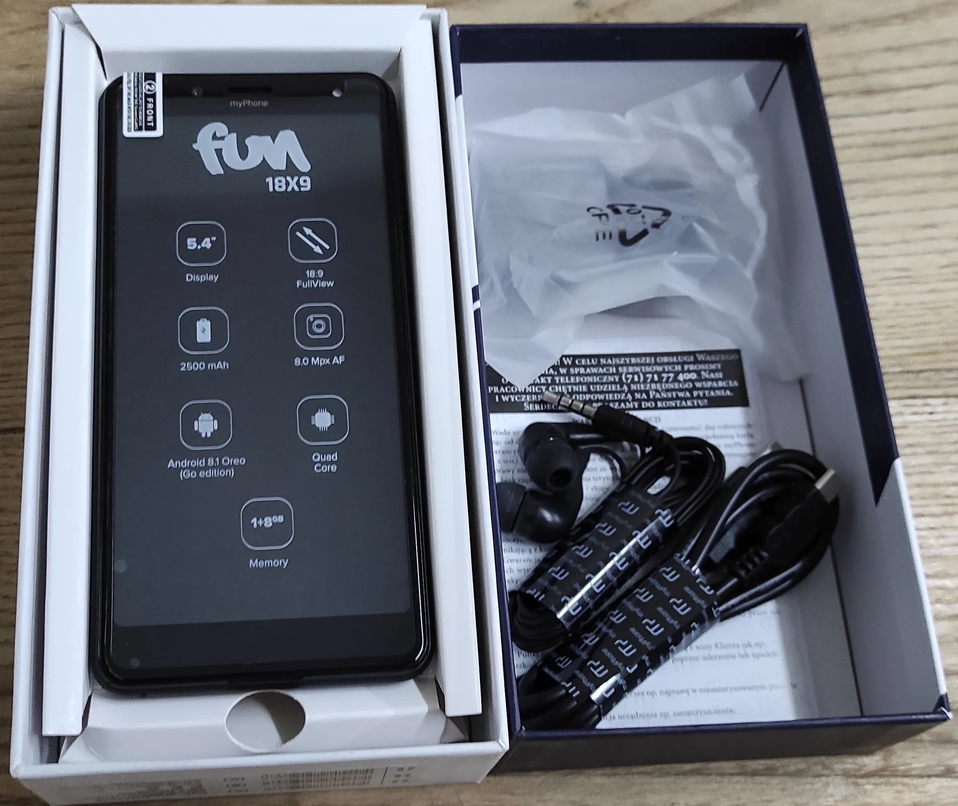 Smartfon marki myPhone w białym pudełku obok ciemnoniebieskiego opakowania z akcesoriami. Na ekranie telefonu widnieje napis 'fun 18X9' oraz specyfikacja techniczna: '5.4" Display', '18:9 FullView', '2500 mAh', '8.0 Mpx AF', 'Android 8.1 Oreo (Go edition)', 'Quad Core' oraz '1+8GB Memory'. W niebieskim pudełku znajdują się czarne słuchawki douszne, kabel USB oraz instrukcja obsługi w języku polskim.
