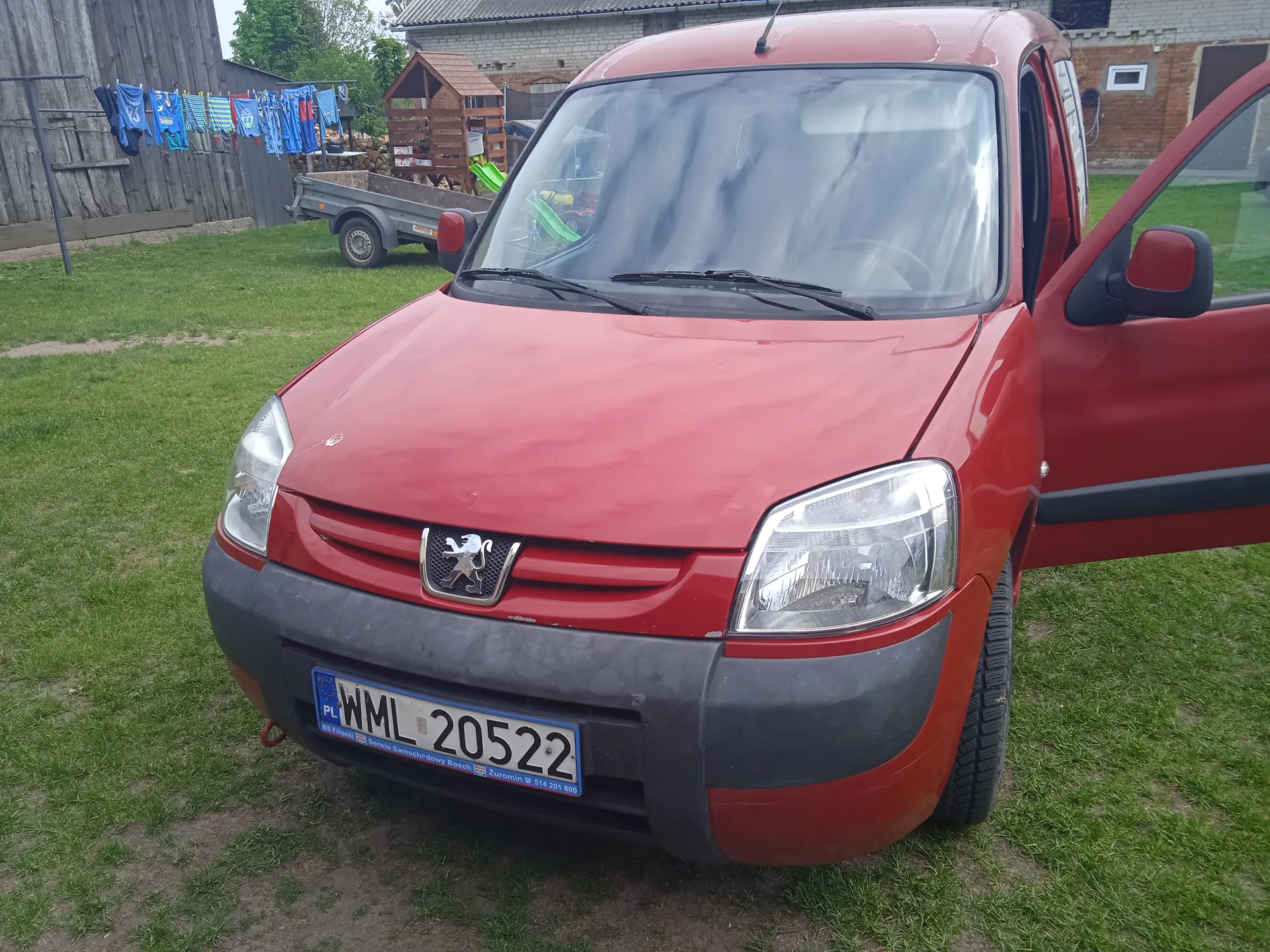 Czerwony samochód dostawczy marki Peugeot z otwartą prawą drzwiami. Widoczna tablica rejestracyjna PL WML 20522 z nieczytelnym tekstem poniżej. Na tle trawa, przyczepa, suszące się niebieskie ubrania na sznurze, drewniana budowla i ceglany budynek. Samochód stoi na trawniku, a w tle widoczne są drzewa i niebo.