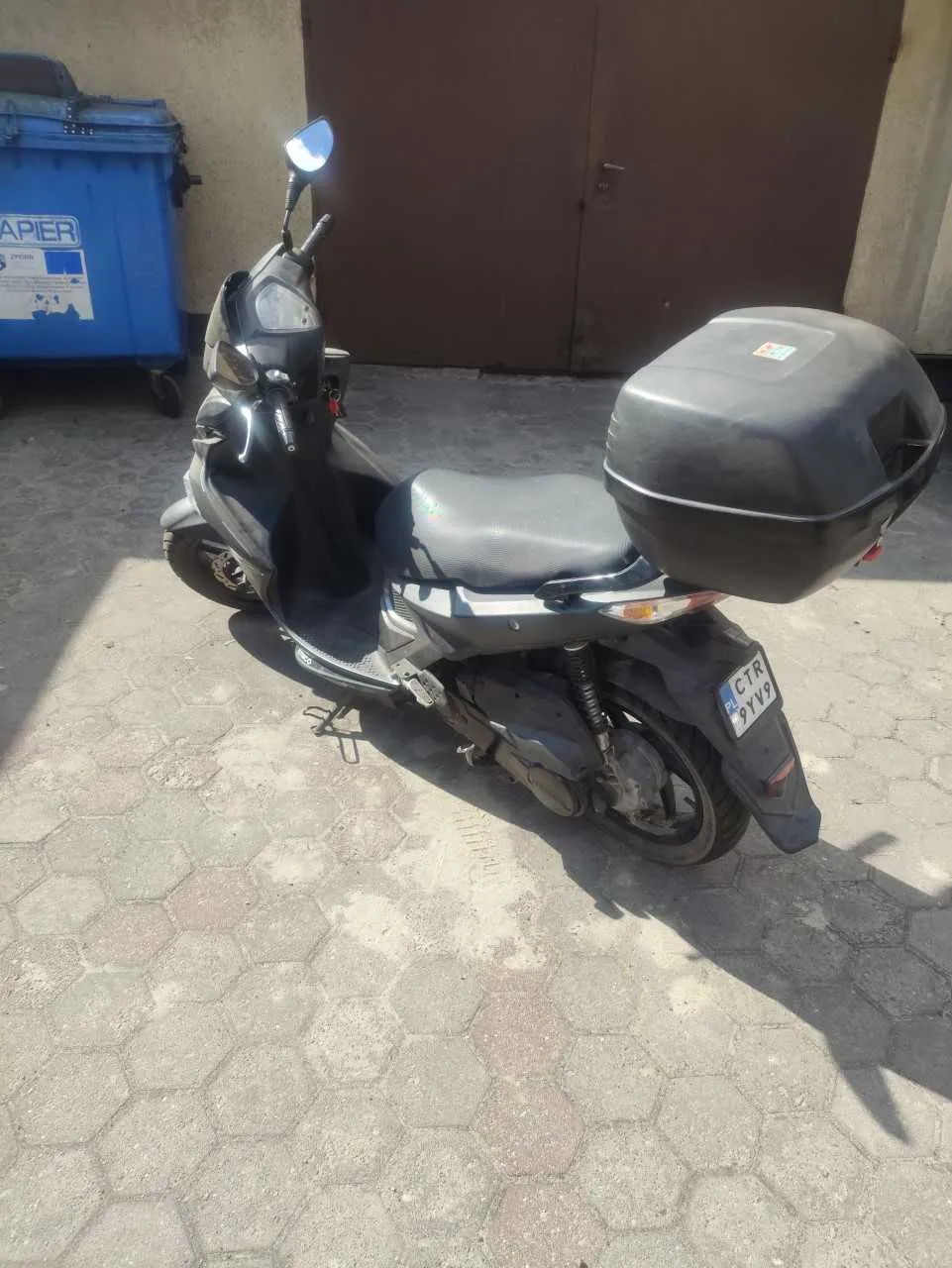 Czarny skuter zaparkowany na chodniku z sześciokątnymi płytkami. Skuter posiada czarny bagażnik na tylnej części, tablicę rejestracyjną z napisem CTR 9YV9 oraz lusterko w lewym górnym rogu. W tle widoczny niebieski pojemnik na śmieci z częściowo widocznym napisem APIER oraz brązowe drzwi garażowe. Oświetlenie wskazuje na słoneczny dzień, cienie padają na powierzchnię chodnika.