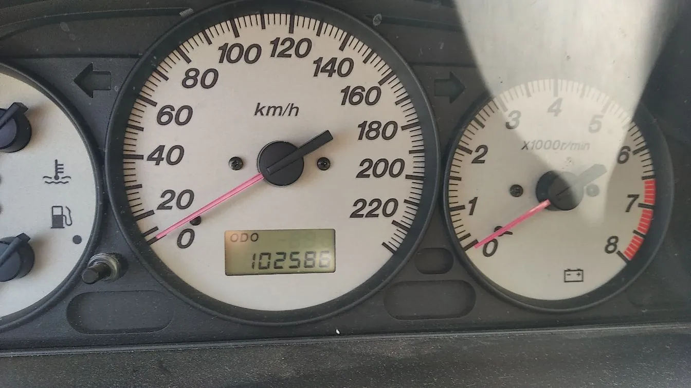 Zbliżenie na deskę rozdzielczą samochodu z dwoma białymi zegarami. Po lewej znajduje się prędkościomierz z oznaczeniem 'km/h', skala sięga 220, a wskazówka wskazuje 0. Poniżej znajduje się cyfrowy licznik przebiegu z tekstem 'ODO 102586'. Po prawej znajduje się obrotomierz z oznaczeniem 'x1000r/min', skala sięga 8, a wskazówka wskazuje 0. Po lewej stronie widoczne są kontrolki temperatury silnika i poziomu paliwa.
