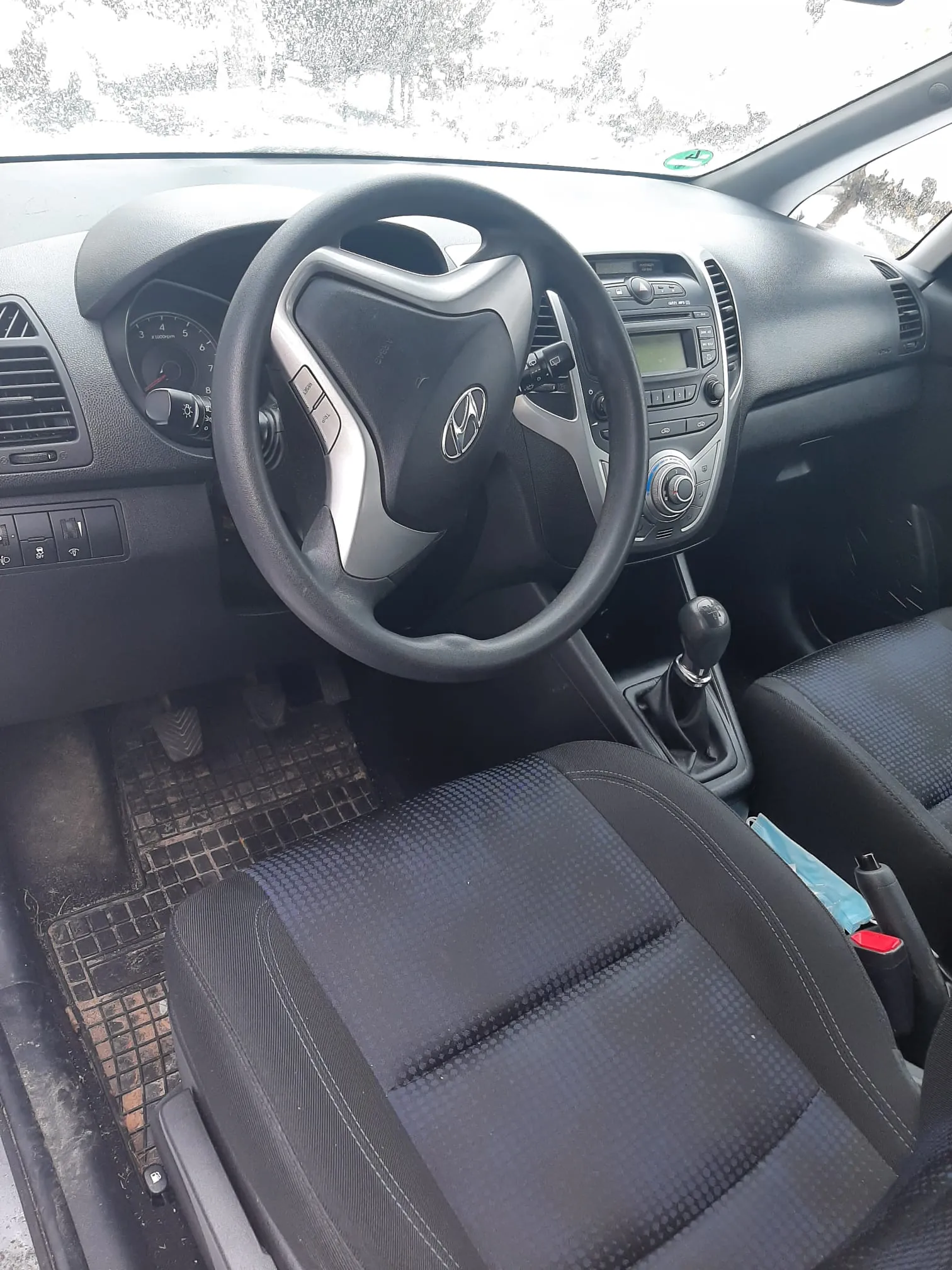 Wnętrze samochodu Hyundai z czarną kierownicą z logo, tablicą rozdzielczą, prędkościomierzem, centralnym panelem z ekranem i przyciskami, czarnymi siedzeniami w wzór, dźwignią biegów oraz brudnymi dywanikami na podłodze. Widok przez przednią szybę na zasneżony krajobraz.