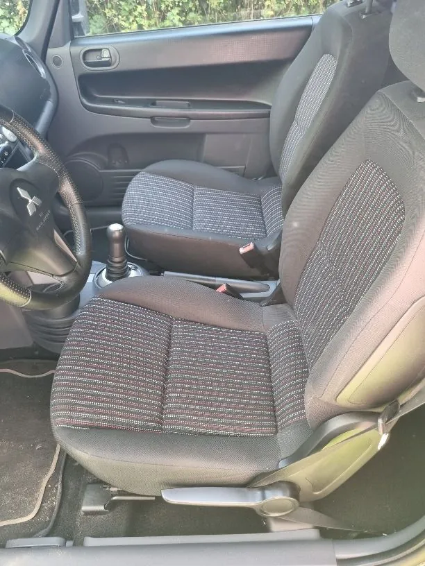 Wnętrze samochodu z widokiem na dwa przednie fotele z ciemnoszarej tkaniny w prążkowany wzór. Po lewej stronie widoczna jest kierownica z logo Mitsubishi oraz lewarek zmiany biegów.