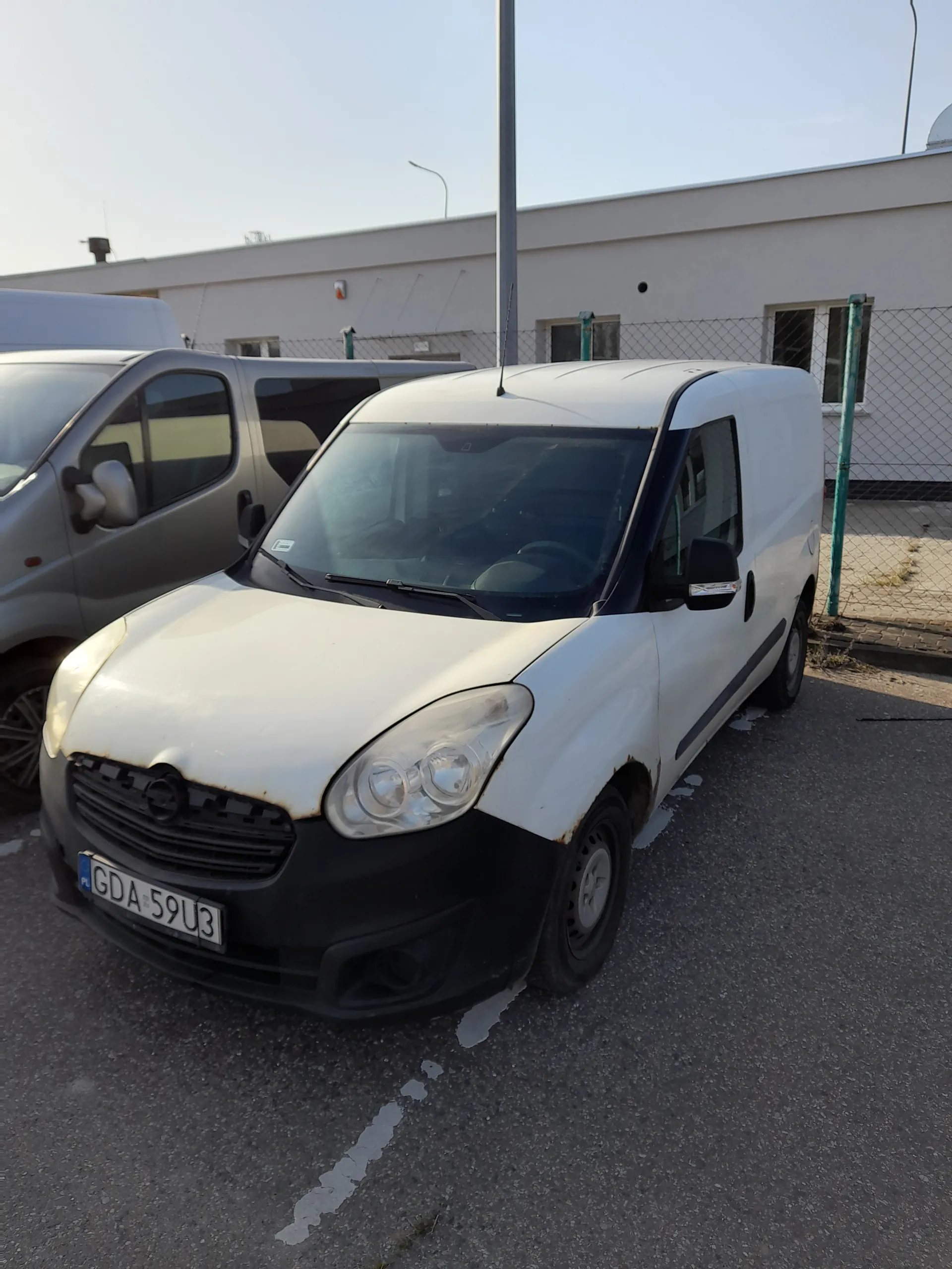 Dwa zaparkowane samochody na asfaltowej powierzchni. Biały samochód z czarnym przodem i tablicą rejestracyjną GDA 59U3 ustawioną pionowo. Szary samochód obok. W tle budynek z siatką ochronną, słupami i jasnym niebem.