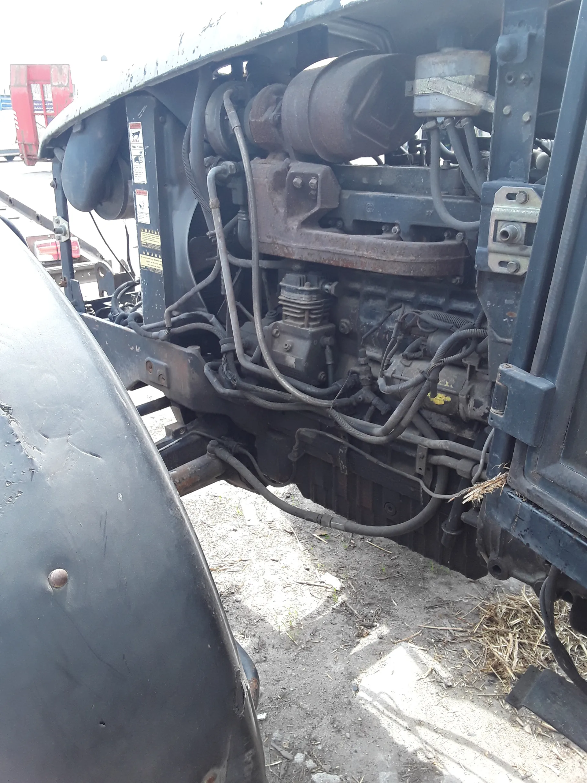 Zbliżenie na komorę silnika pojazdu, zawierającą liczne przewody, metalowe elementy i części mechaniczne. Widoczne są ślady korozji na niektórych elementach. Po prawej stronie obrazu znajduje się ziemia z suchą trawą i drobnymi kamieniami. Na jednym z elementów umieszczono etykietę z ostrzeżeniami i symbolami.