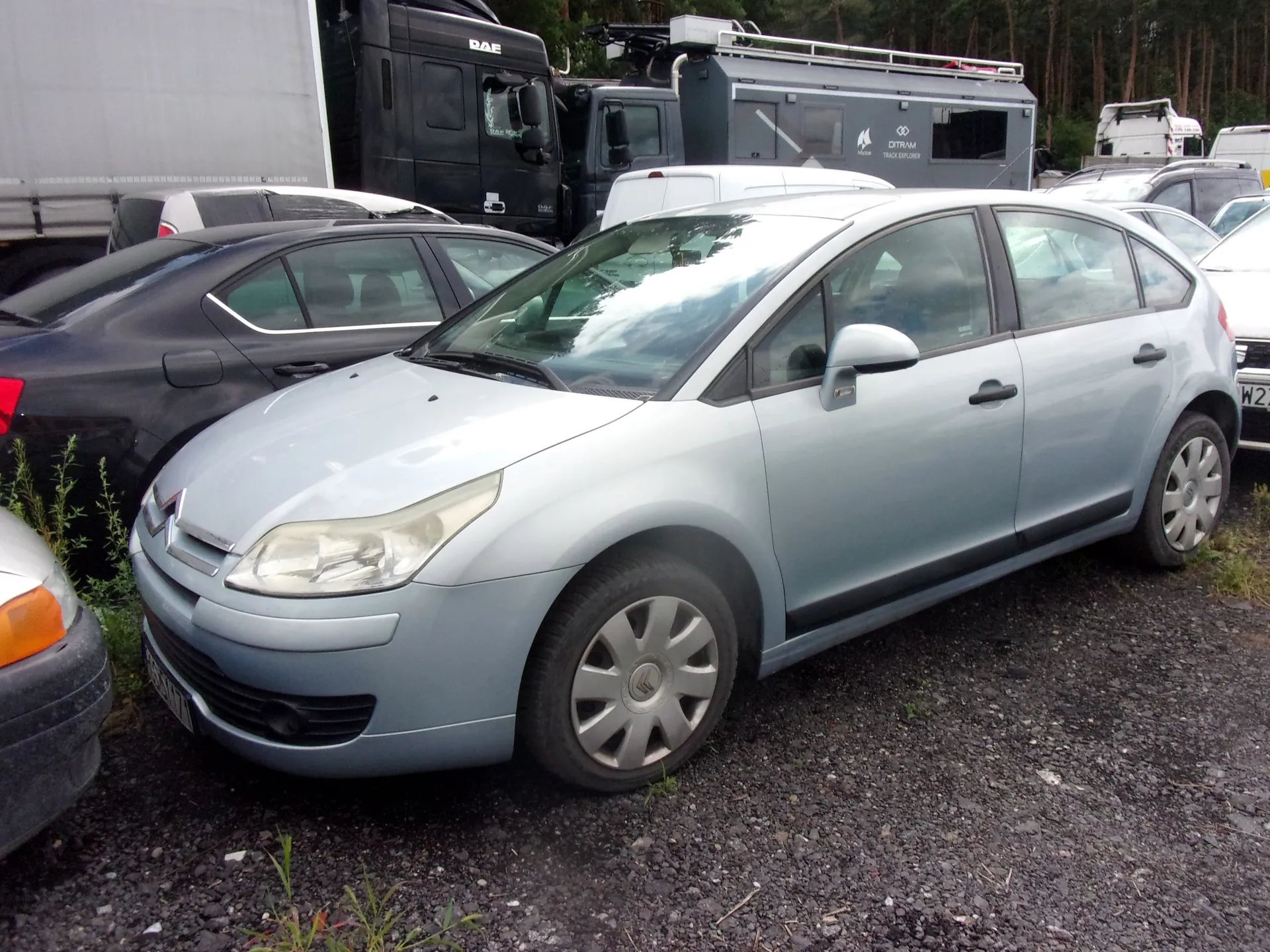 Jasnoniebieski samochód osobowy marki Citroën zaparkowany na żwirowym placu. Auto stoi bokiem do obiektywu, ukazując przód i prawy profil. W tle widoczne są inne zaparkowane samochody, biały kamper oraz czarna ciężarówka marki DAF.