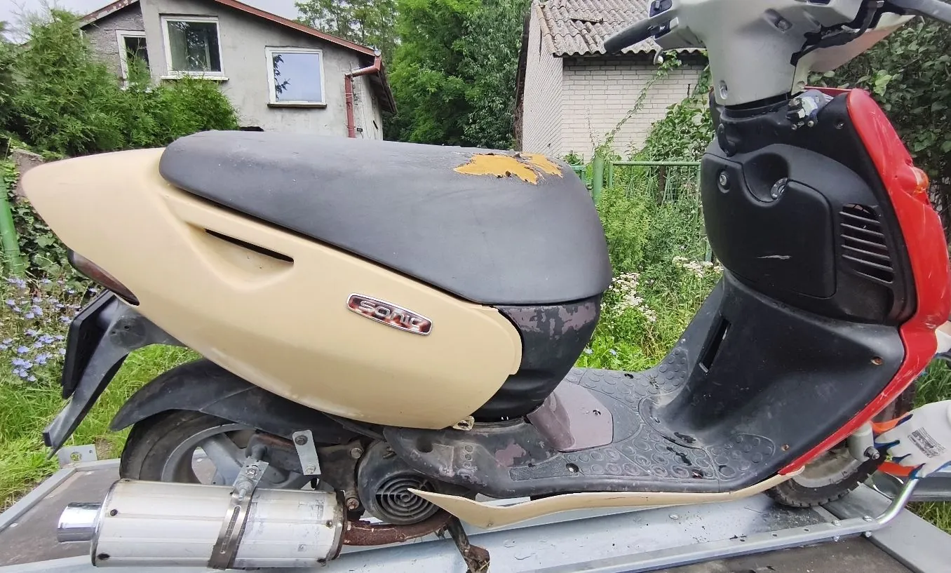 Zdjęcie przedstawiające skuter w kolorach beżowym i czerwonym, zaparkowany na metalowej platformie. Pojazd ma czarne siedzenie z widocznym uszkodzeniem materiału na górnej części. Na boku beżowej owiewki znajduje się napis 'Sonic'. W tle widoczne są dwa budynki mieszkalne oraz zielona roślinność i ogrodzenie.