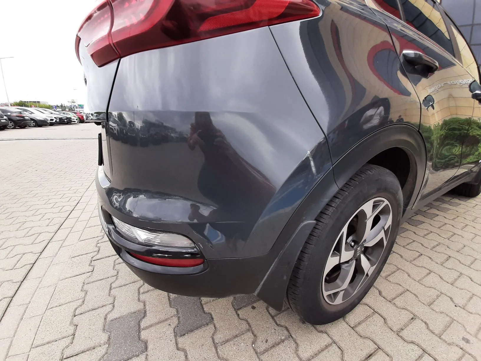 Zbliżenie na tylny prawy błotnik i lampę samochodu w kolorze ciemnoszarym. Na karoserii widoczne są zarysowania oraz wgniecenia. Samochód stoi na parkingowej nawierzchni z kostki brukowej.
