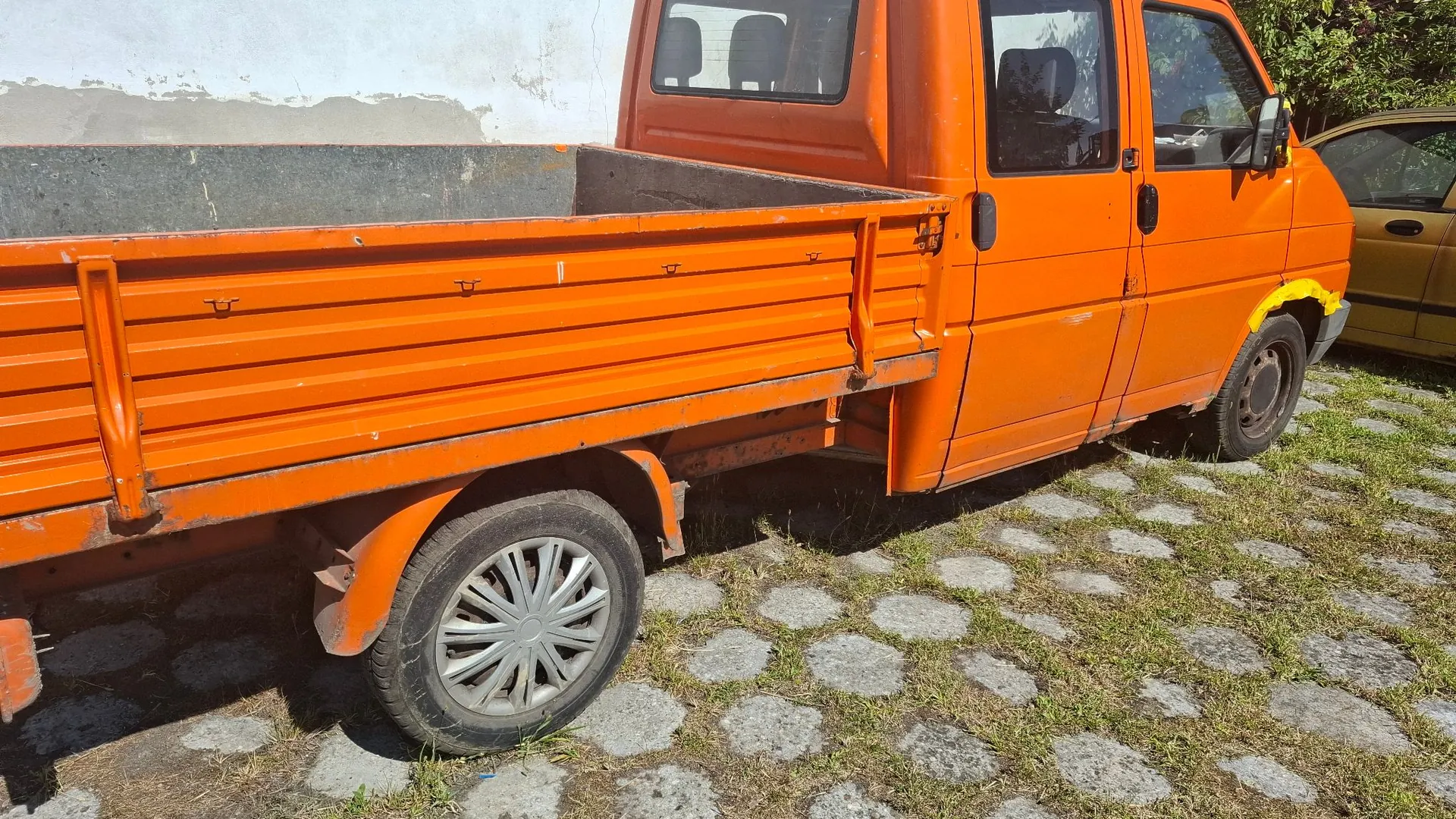 Pomarańczowy samochód dostawczy typu pick-up zaparkowany na podjeździe z kostki brukowej z trawą pomiędzy kamieniami. Widoczna jest boczna część pojazdu z otwartą skrzynią ładunkową oraz fragment drugiego, żółtego samochodu w tle po prawej stronie.