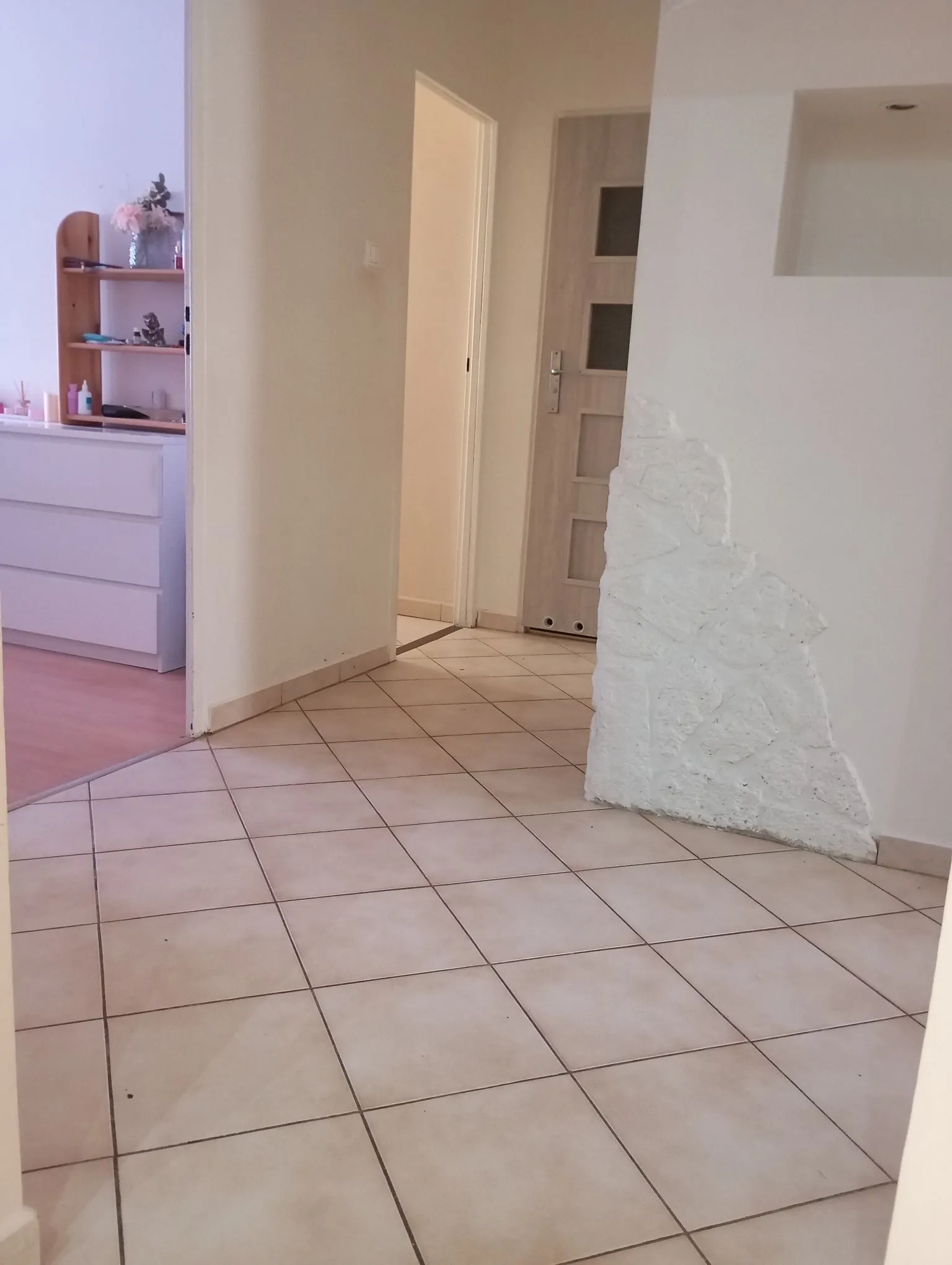 Wnętrze jasnego pomieszczenia z kafelkami w kolorze beżowym na podłodze. Po prawej stronie znajduje się biała ściana z fragmentem odsłoniętego, pomalowanego na biało kamienia lub tynku o nieregularnej strukturze. W głębi widoczne są otwarte drzwi prowadzące do innego pokoju oraz zamknięte białe drzwi wewnętrzne z przeszkleniami. Po lewej stronie widać fragment białej komody oraz drewniany regał z ozdobami.