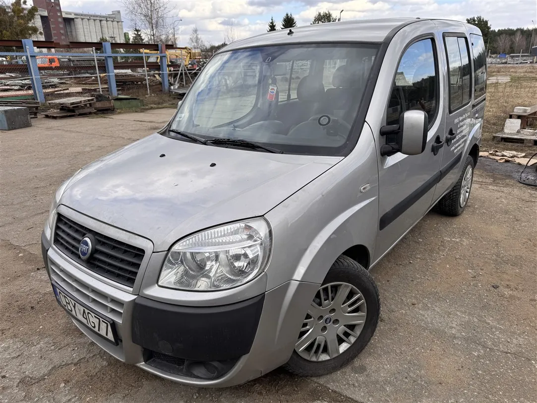 Srebrny samochód dostawczy marki Fiat Doblo zaparkowany na piaszczystym placu. Pojazd jest widoczny z przodu i z boku. Na przednim zderzaku znajduje się tablica rejestracyjna o numerze CBY 4G77. W tle widoczne są elementy infrastruktury przemysłowej, ogrodzenie oraz budynki.
