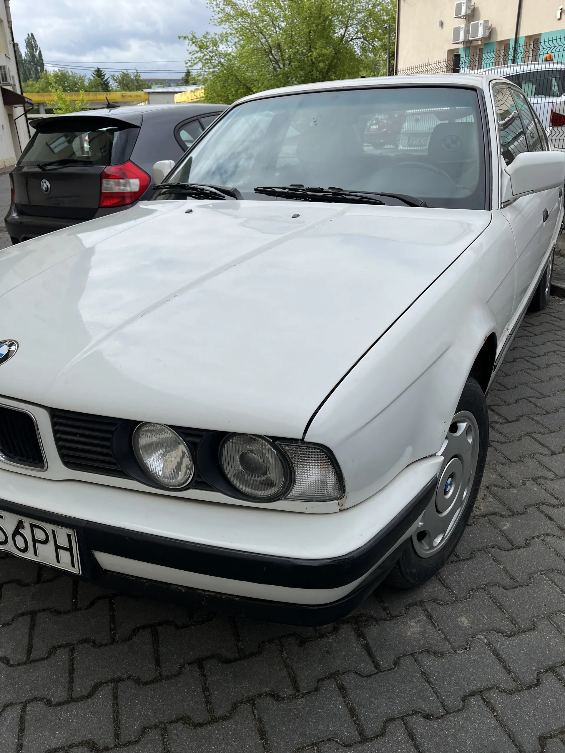 Biały samochód marki BMW, widok z przodu, trzy okrągłe reflektory, tablica rejestracyjna z napisem "56PH". Samochód zaparkowany na brukowanej powierzchni. W tle drugi, ciemny samochód, budynki, drzewa oraz częściowo zachmurzone niebo.