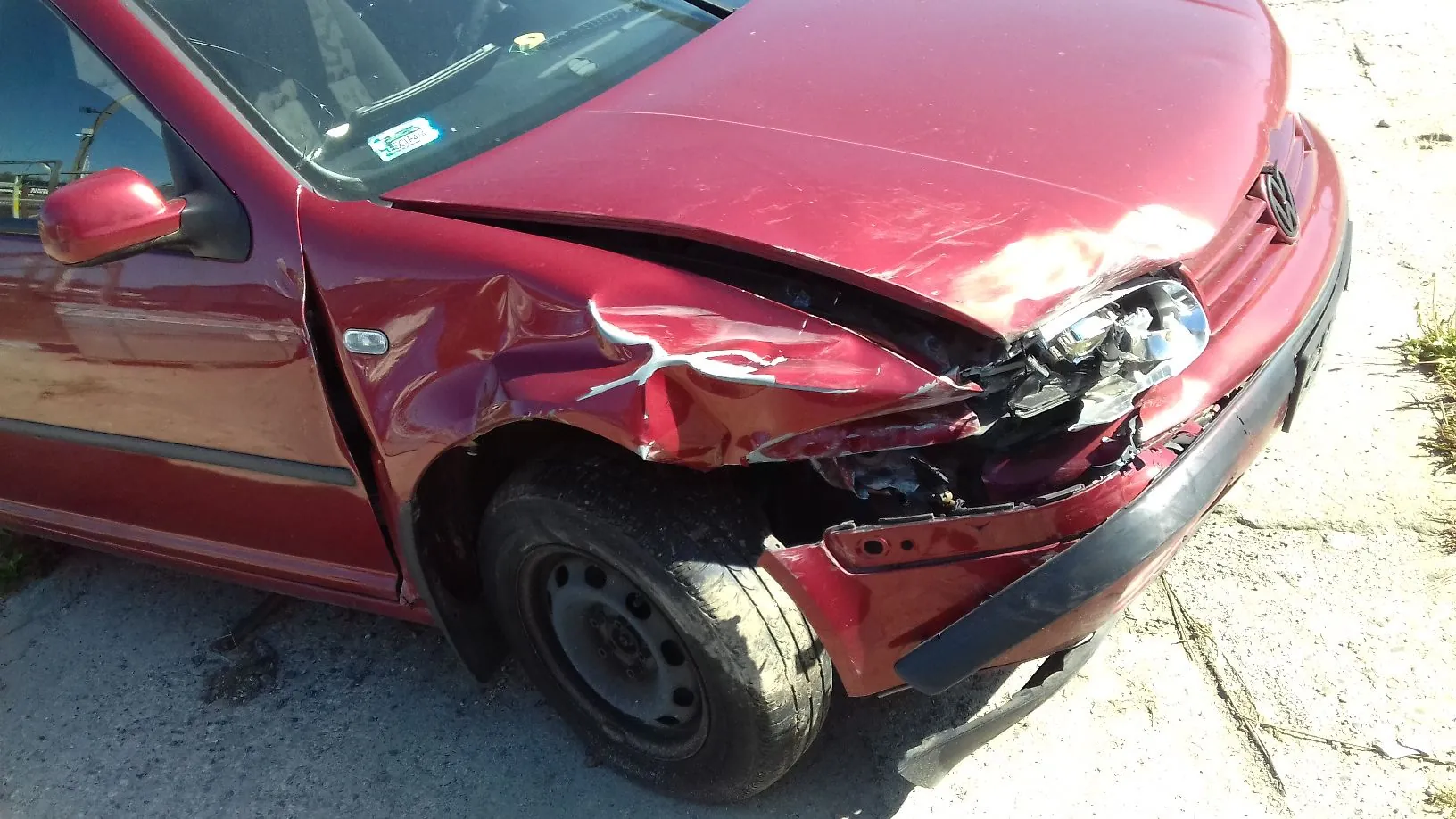 Zbliżenie na przód bordowego samochodu osobowej marki Volkswagen, który posiada znaczne uszkodzenia karoserii po prawej stronie. Widoczne jest wgniecenie błotnika, zniszczony reflektor oraz odkształcona maska i zderzak. Samochód stoi na piaszczystym podłożu.