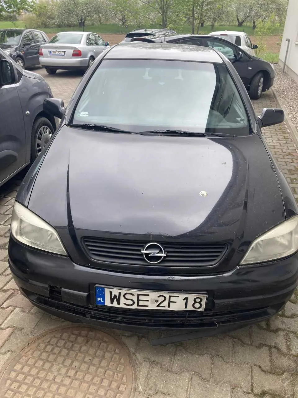 Czarny samochód marki Opel z tablicą rejestracyjną WSE 2F18 (PL) zaparkowany na brukowanym placu. W tle widoczne są inne pojazdy, drzewa oraz budynek po prawej stronie. Na masce samochodu znajduje się logo Opel, a przednia szyba jest przejrzysta. Pod samochodem widoczna klapa kanalizacyjna.