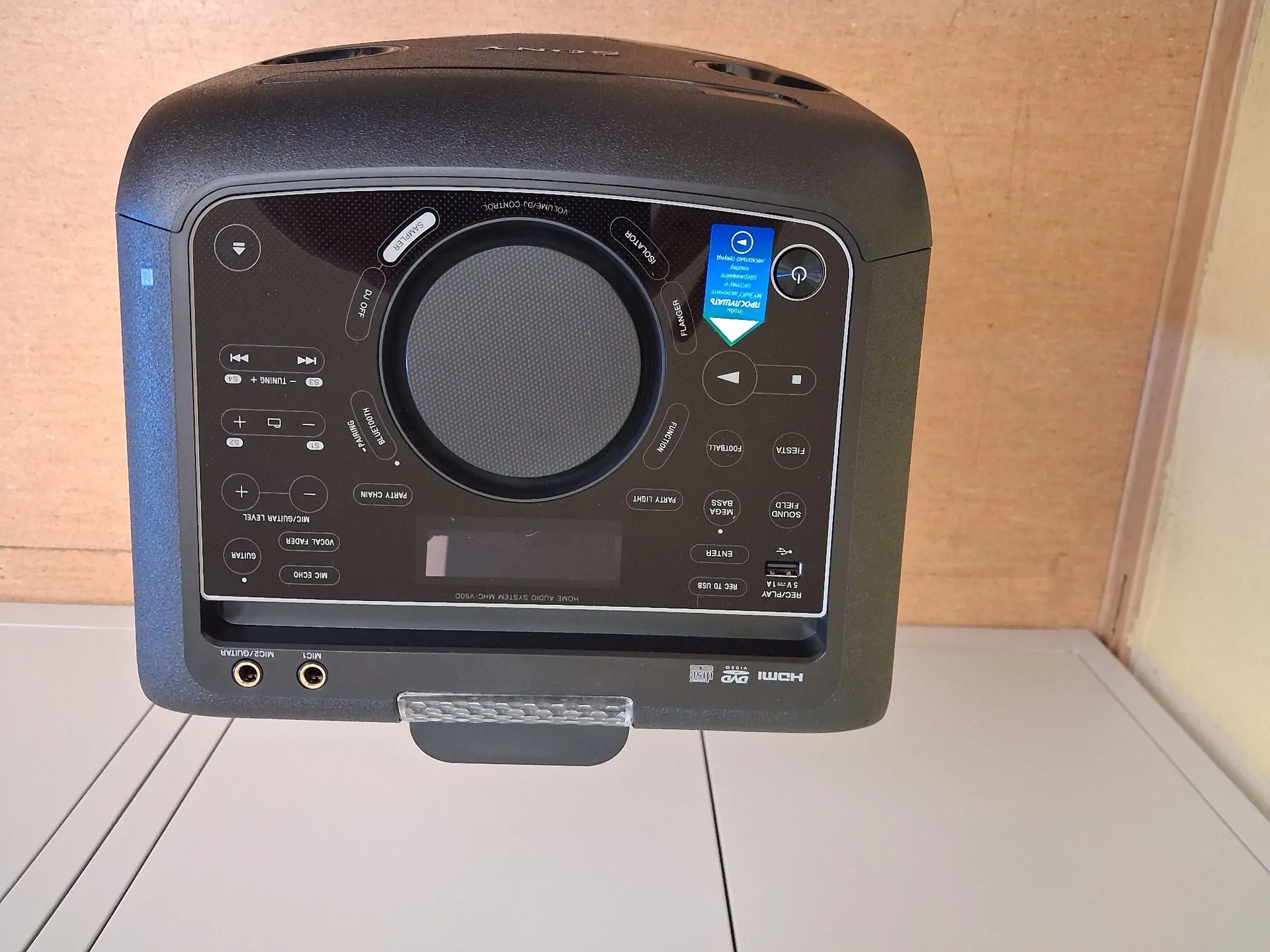 Czarny system audio marki LG z panelem sterowania na górze. Na panelu znajdują się liczne przyciski funkcyjne, pokrętło głośności oraz wyświetlacz. Widoczne napisy obejmują m.in. 'HDMI', 'DVD', 'USB', 'BLUETOOTH', 'MIC ECHO' oraz 'GUITAR'. Na jednym z przycisków znajduje się niebieska naklejka z napisem 'PRODUKTY'. Urządzenie posiada uchwyt po prawej stronie.