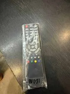 Pilot zdalnego sterowania owinięty w przezroczystą folię. Na pilocie znajdują się przyciski, w tym okrągły pad nawigacyjny oraz kolorowe przyciski (czerwony, zielony, żółty, niebieski). Na folii widoczny napis '150.00'. Pilot leży na ciemnej powierzchni.