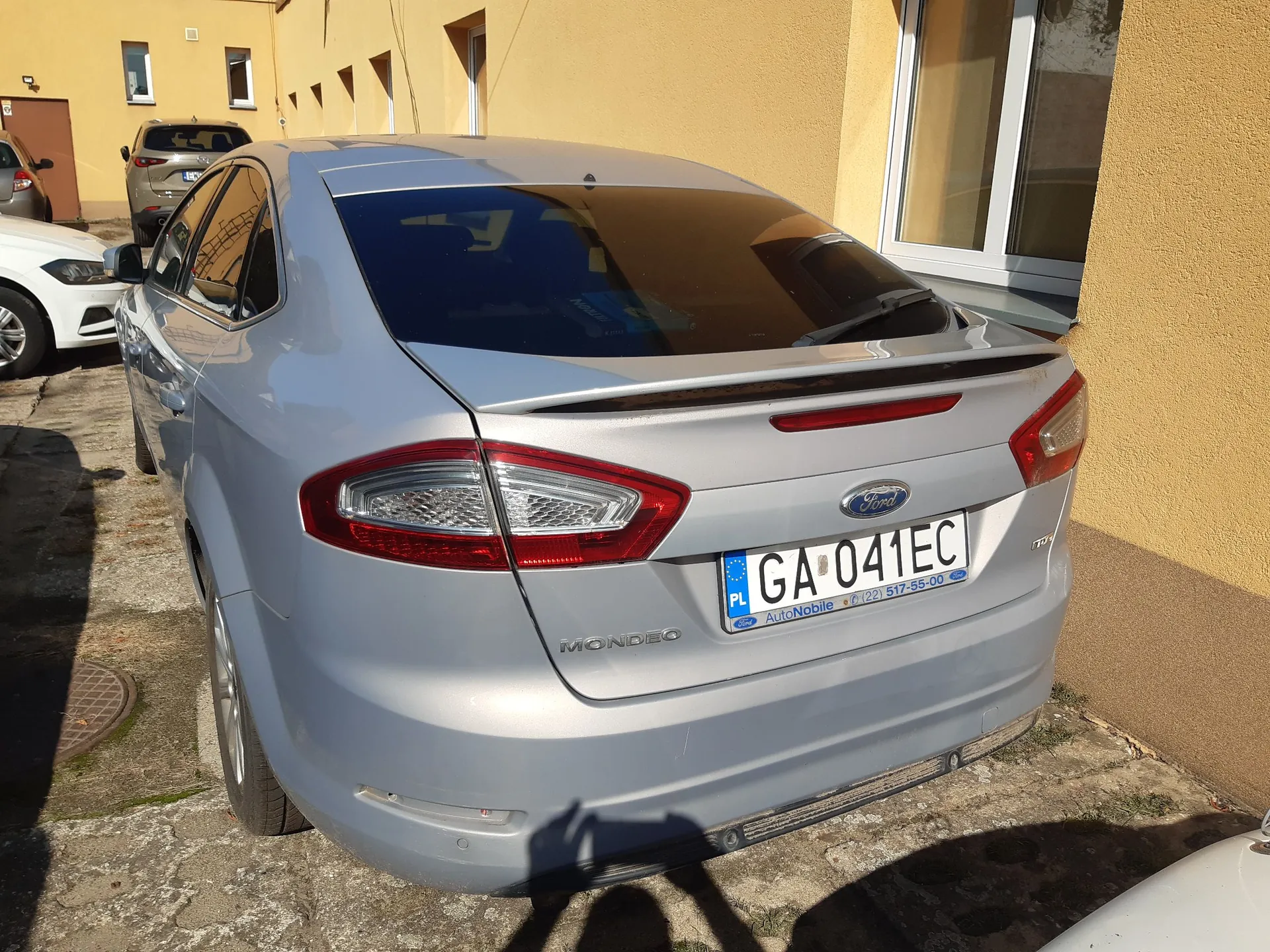 Srebrny samochód marki Ford Mondeo zaparkowany tyłem do żółtej ściany budynku. Widoczna jest tylna część pojazdu z tablicą rejestracyjną o numerze GA 041EC. Na tablicy znajduje się również napis 'AutoNoble' oraz numer telefonu '01221 517-55-00'. W tle widoczny jest fragment innego białego samochodu oraz okna budynku.
