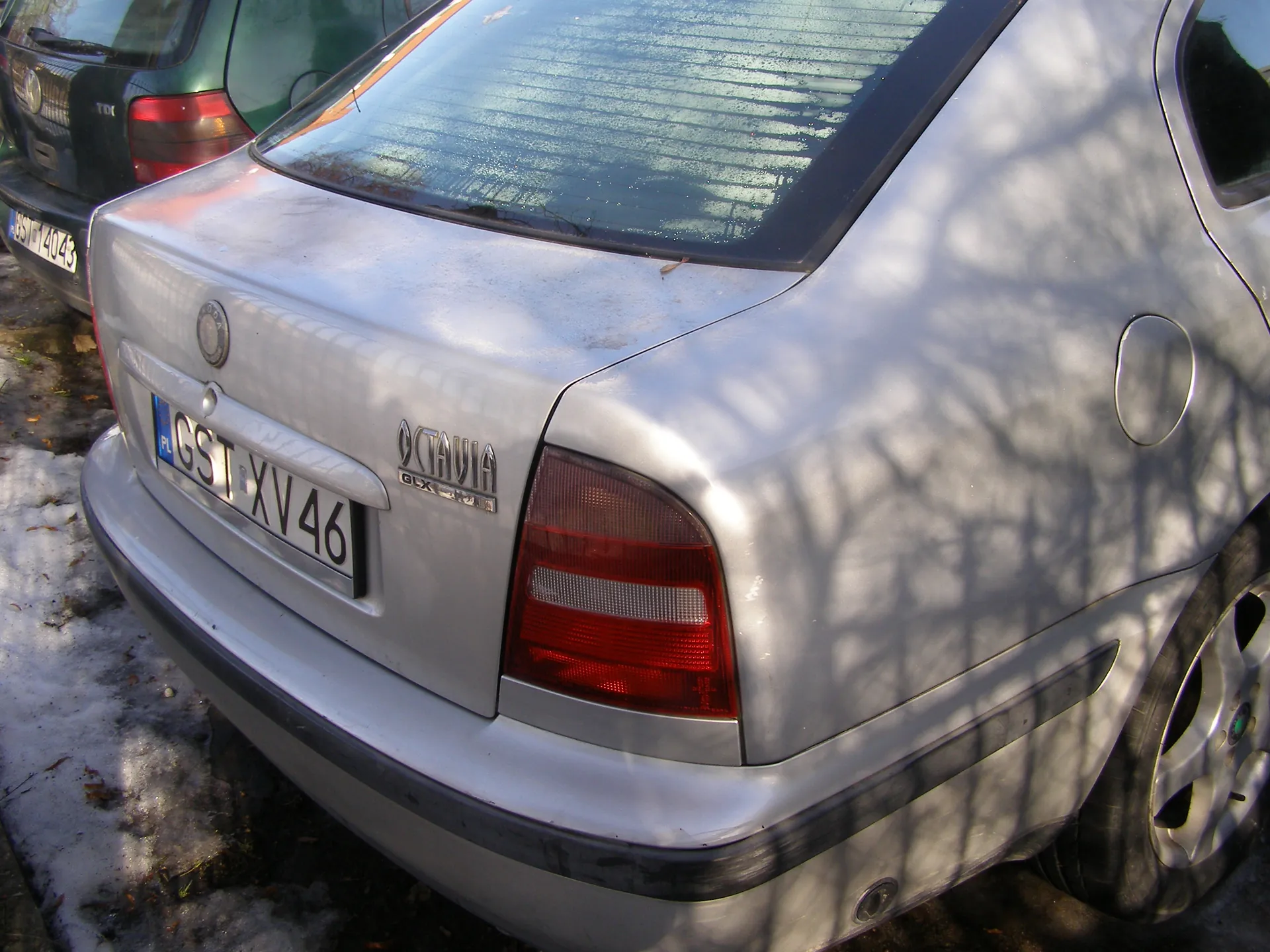 Zbliżenie na tył srebrnego samochodu marki Škoda Octavia GLX z tablicą rejestracyjną PL GST XV 46. Widoczne są czerwone światła tylnego, logo marki oraz napis "OCTAVIA GLX". W tle częściowo widoczny zielony samochód z tablicą GST 4043. Na ziemi resztki śniegu i liście, na karoserii cienie drzew.