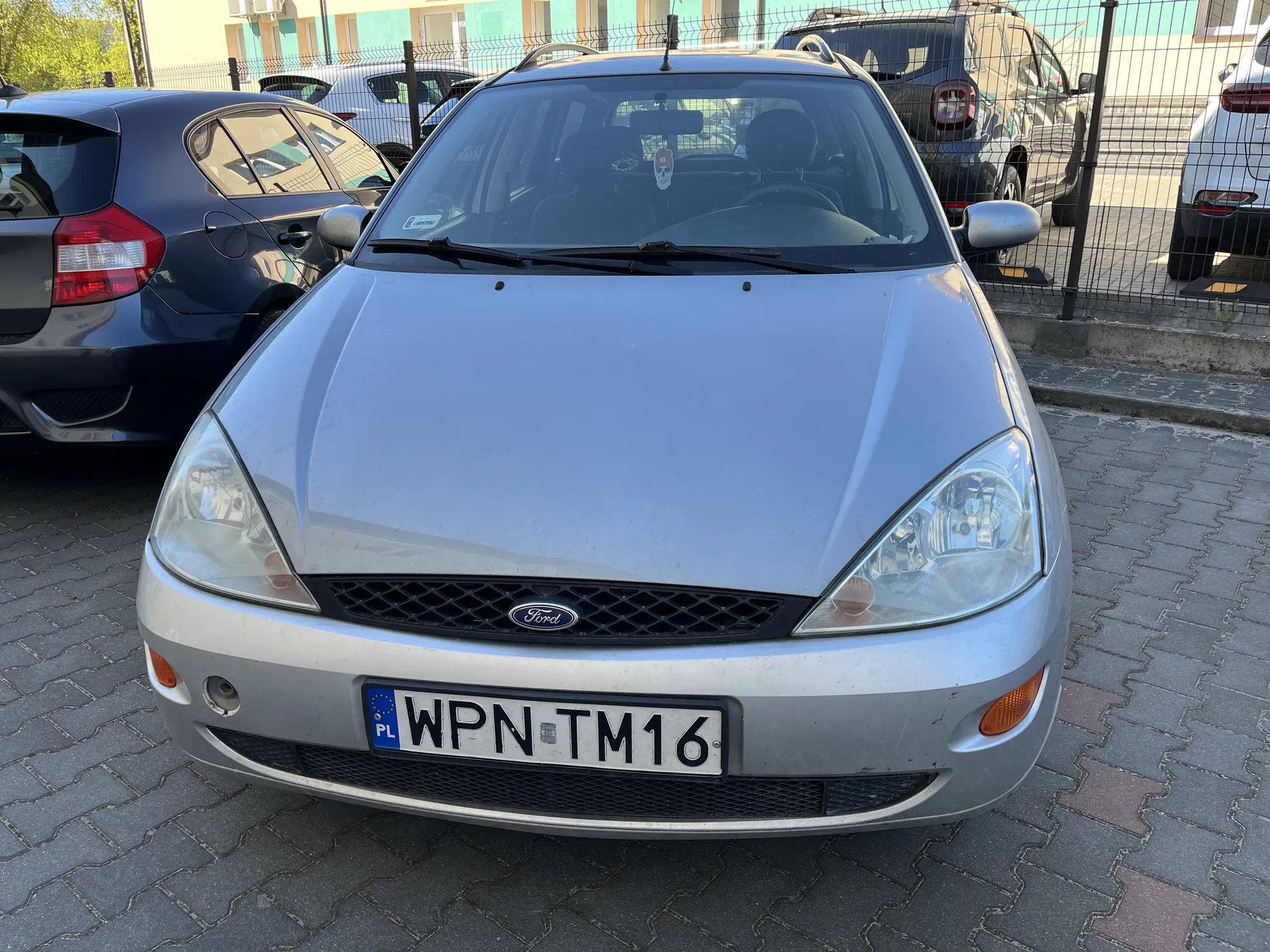 Srebrny samochód marki Ford z tablicą rejestracyjną WPN TM16 (z niebieskim pasem z napisem PL po lewej stronie) zaparkowany na brukowanym placu. W tle widoczne są inne pojazdy, metalowe ogrodzenie oraz budynek z kolorowymi oknami. Na przedniej szybie samochodu widoczne są przedmioty wiszące na lusterku wstecznym.